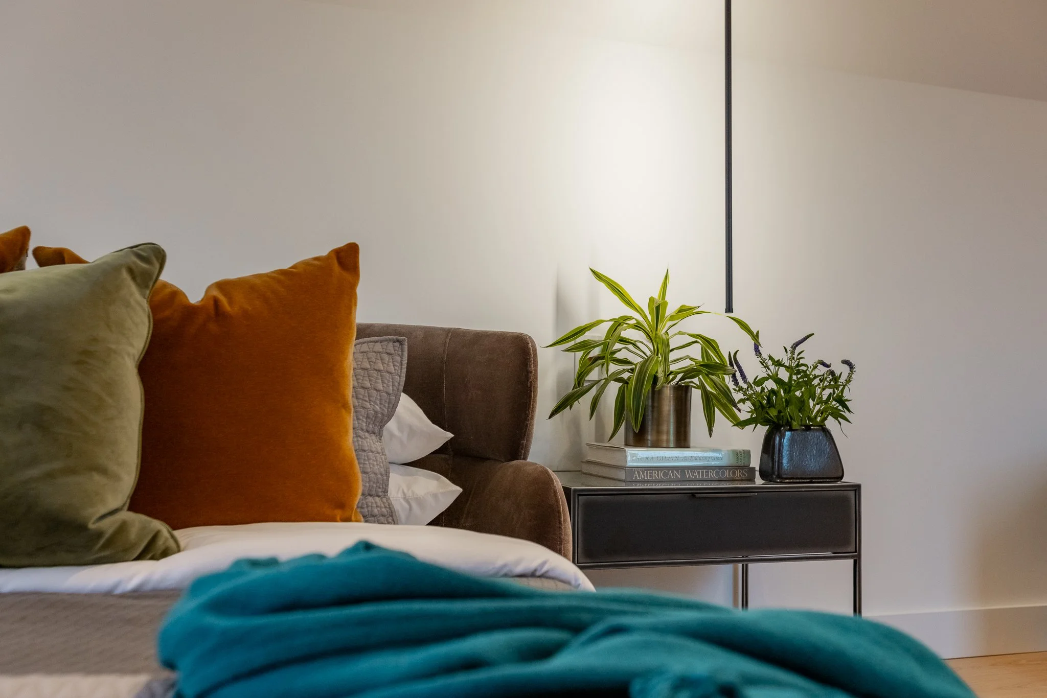 Living room with a bed, colorful pillows, a black bedside table with potted plants, books, and a white wall.
