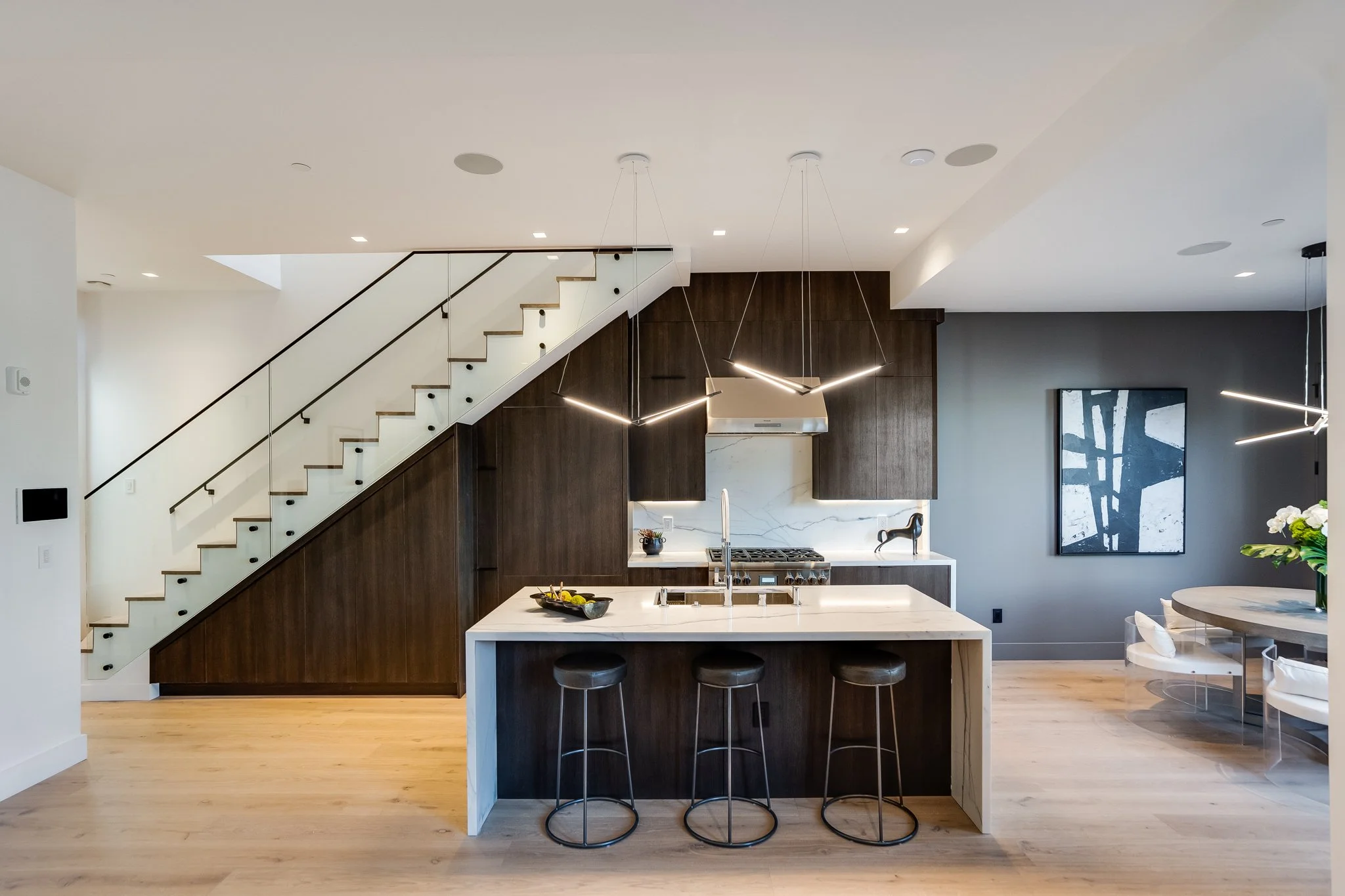 Modern kitchen with dark wood cabinetry, white marble backsplash, island with seating, staircase with glass railing, and contemporary lighting fixtures.