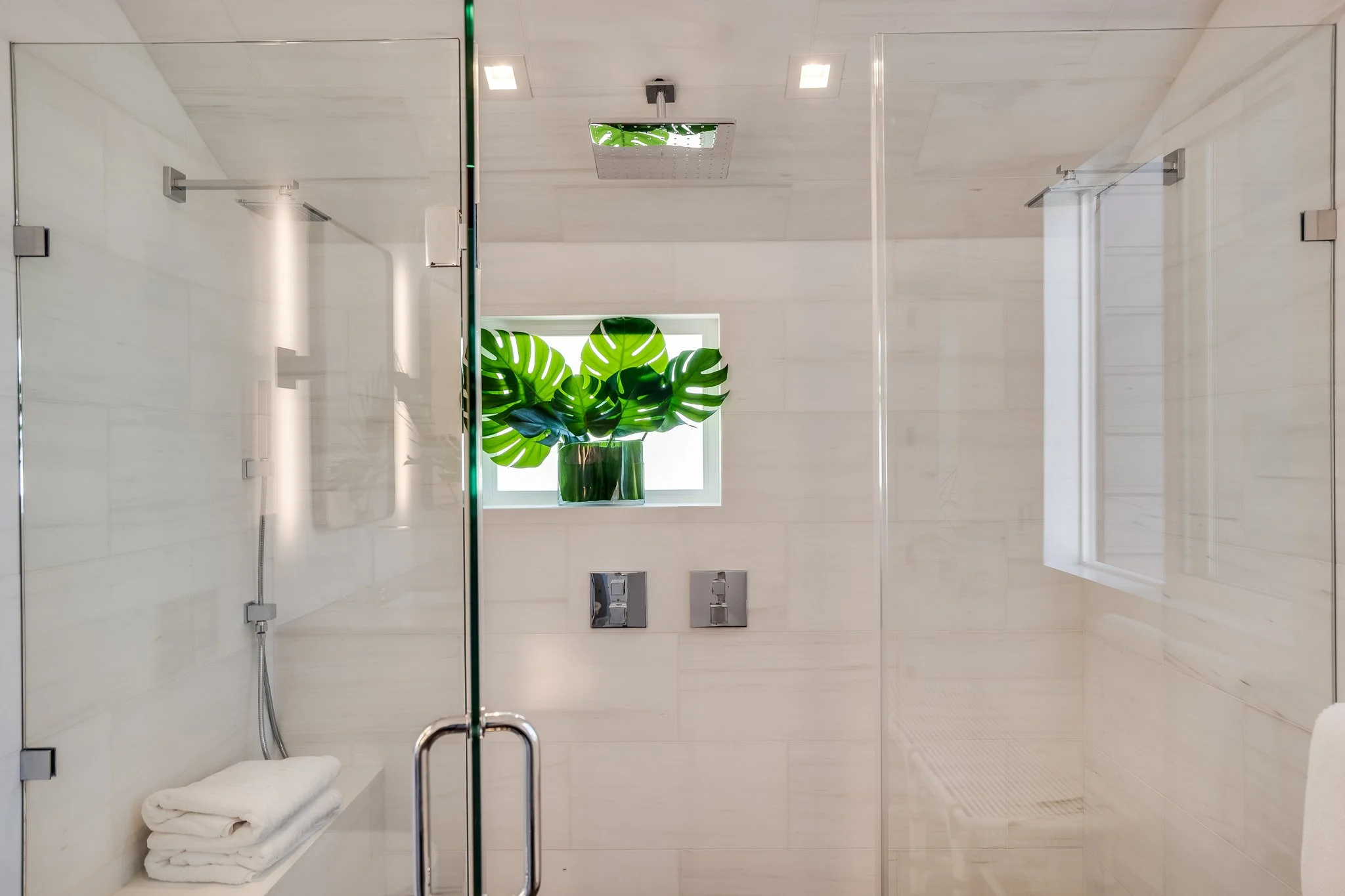Modern bathroom with beige tiled walls, a glass shower enclosure, a window with a green monstera plant in a glass vase, and neatly folded white towels.