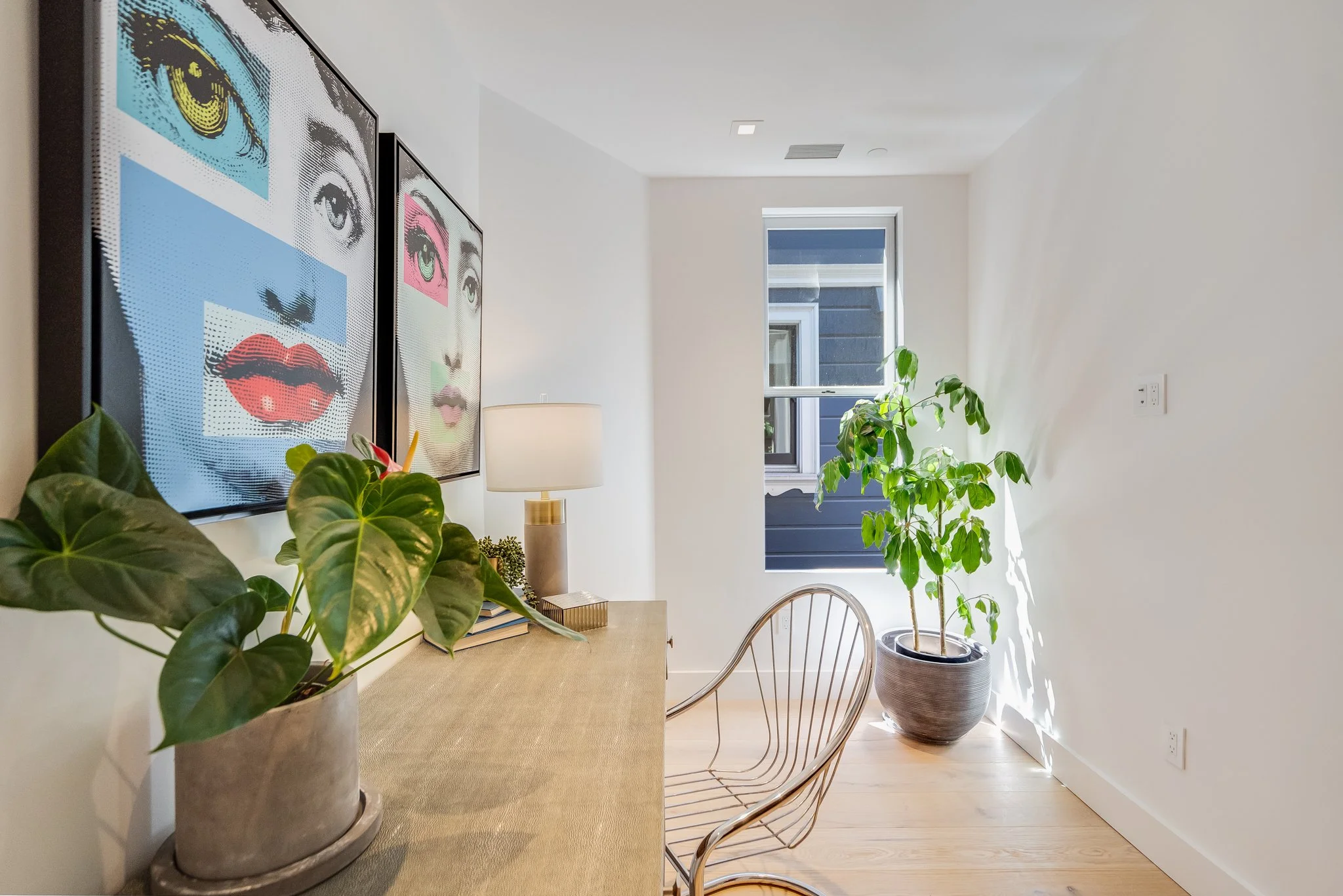 Bright living room corner with wall art, a table with a potted plant, a table lamp, a wire chair, and a window with sunlight streaming in.