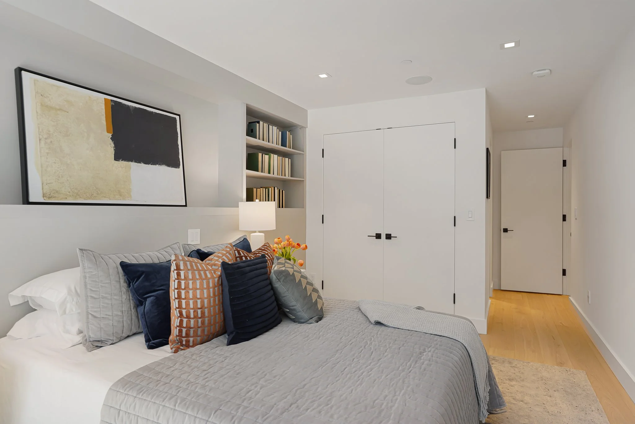A modern bedroom with white walls and ceiling, a bed with a light gray quilt and multiple pillows in shades of gray, blue, and orange, a nightstand with a white lamp and vase of orange tulips, a built-in bookshelf with books, abstract artwork on the 
