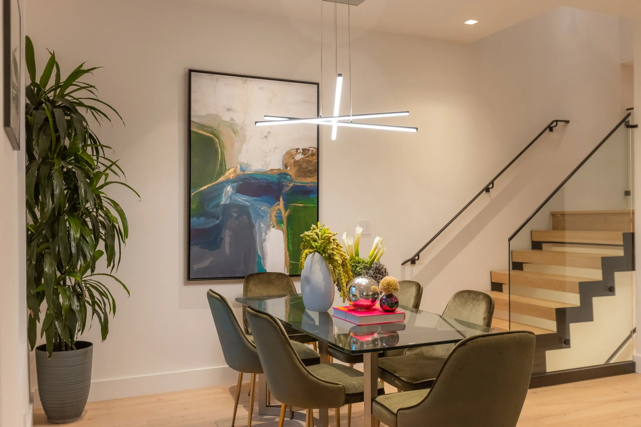 Modern dining room with six green upholstered chairs around a glass table, a large abstract painting on the wall, a potted plant, and a staircase in the background. Artful chandelier hanging above the table.