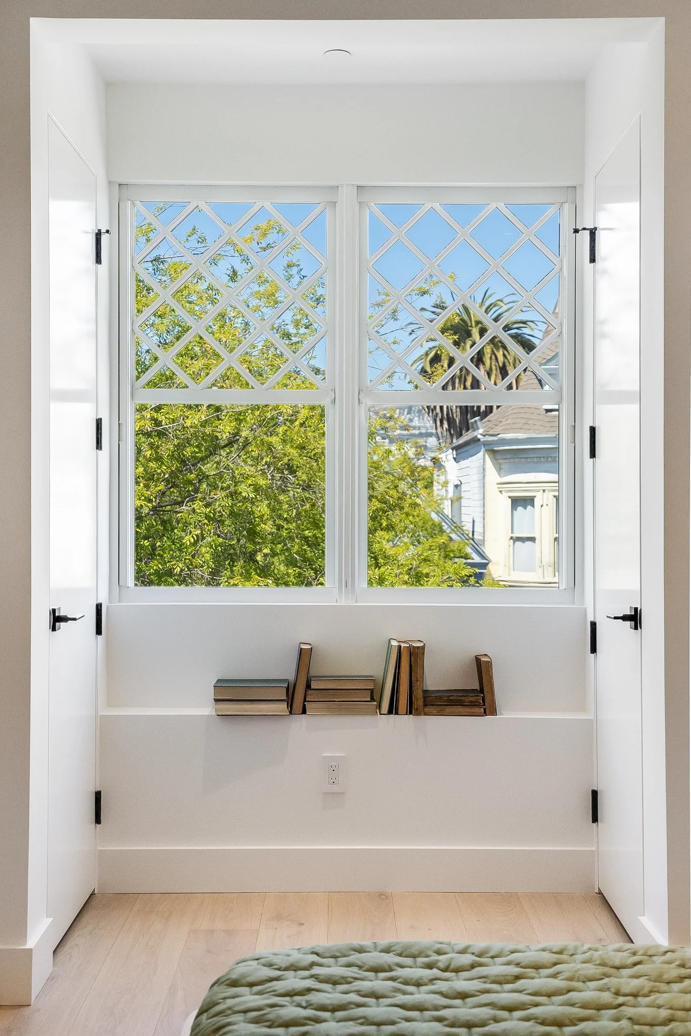 A window with diamond lattice pattern on the upper pane and two lower clear panes showing trees and houses outside. White walls and wood flooring, with a shelf holding stacked books beneath the window.