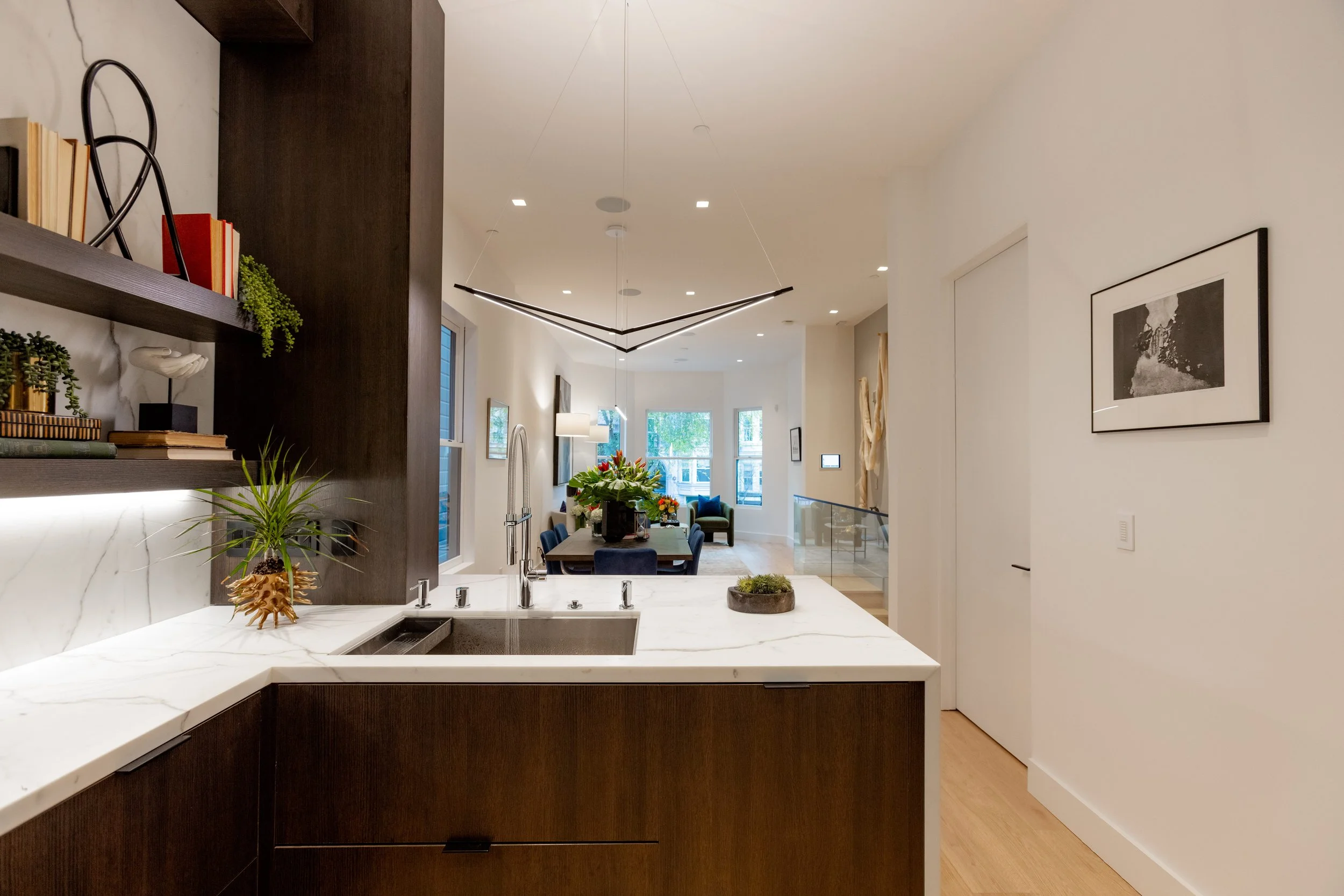 Modern kitchen with white marble countertops, dark wood cabinetry, and decorative shelving with books and plants, overlooking a dining and living area with large windows and contemporary furniture.