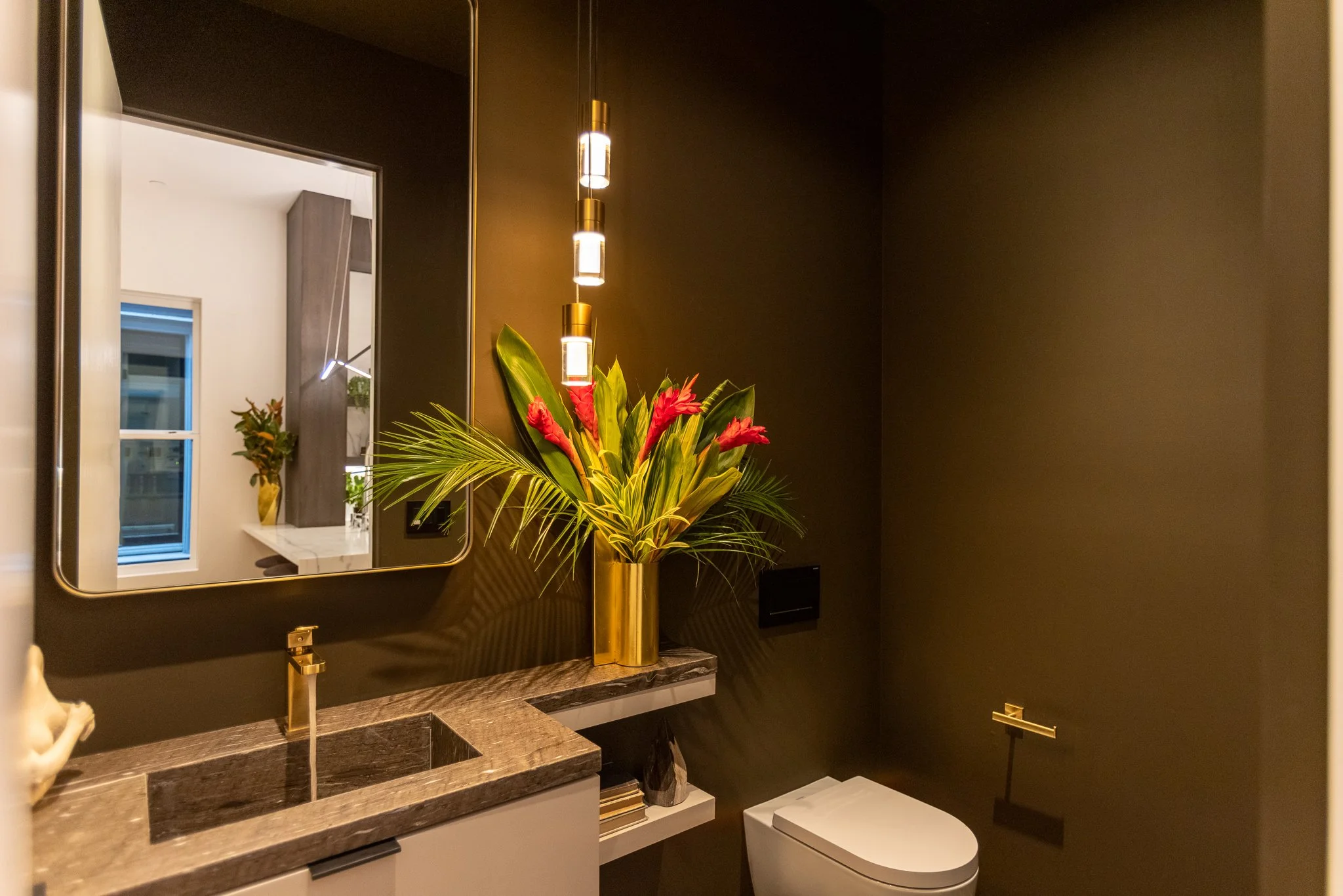 Modern bathroom with dark walls, gold fixtures, a large mirror, a marble countertop with a built-in sink, a tall vase with pink and green plants, and a toilet.