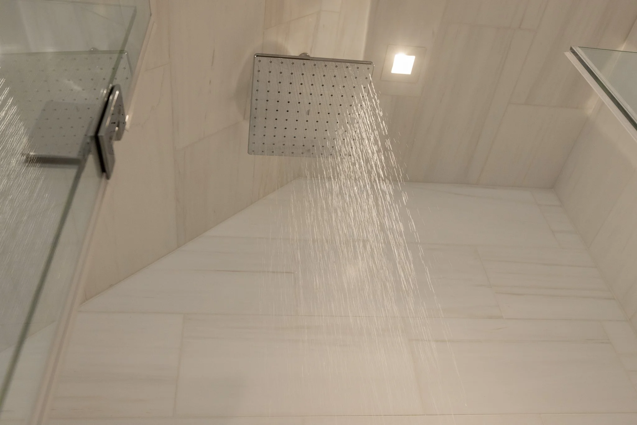 A modern shower with a square rainfall showerhead installed on a tiled ceiling, water flowing from the showerhead, with a glass shower door on the left and a recessed ceiling light.