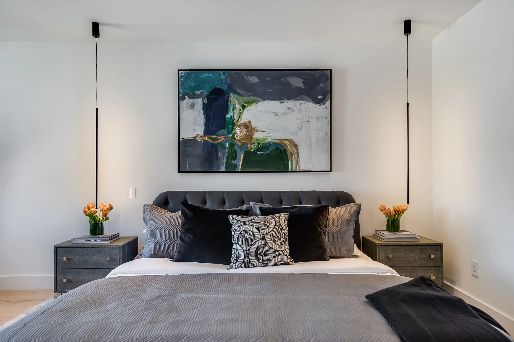 A modern bedroom with a dark gray upholstered bed, black decorative pillows, and bedding in neutral tones. Two matching gray nightstands with stacks of books and orange tulip flowers in glass vases. Abstract artwork hangs above the bed, and two penda
