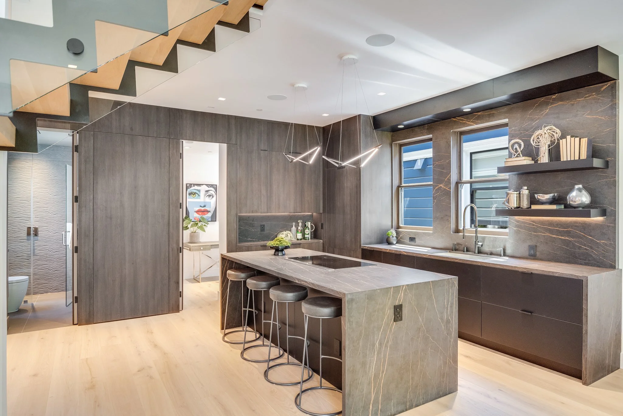 Modern kitchen with gray marble countertops and backsplash, dark wood cabinets, three barstools at the island, two windows above the sink, decorative shelves with books and decorative items, an abstract art piece in a nearby room, and a staircase wit