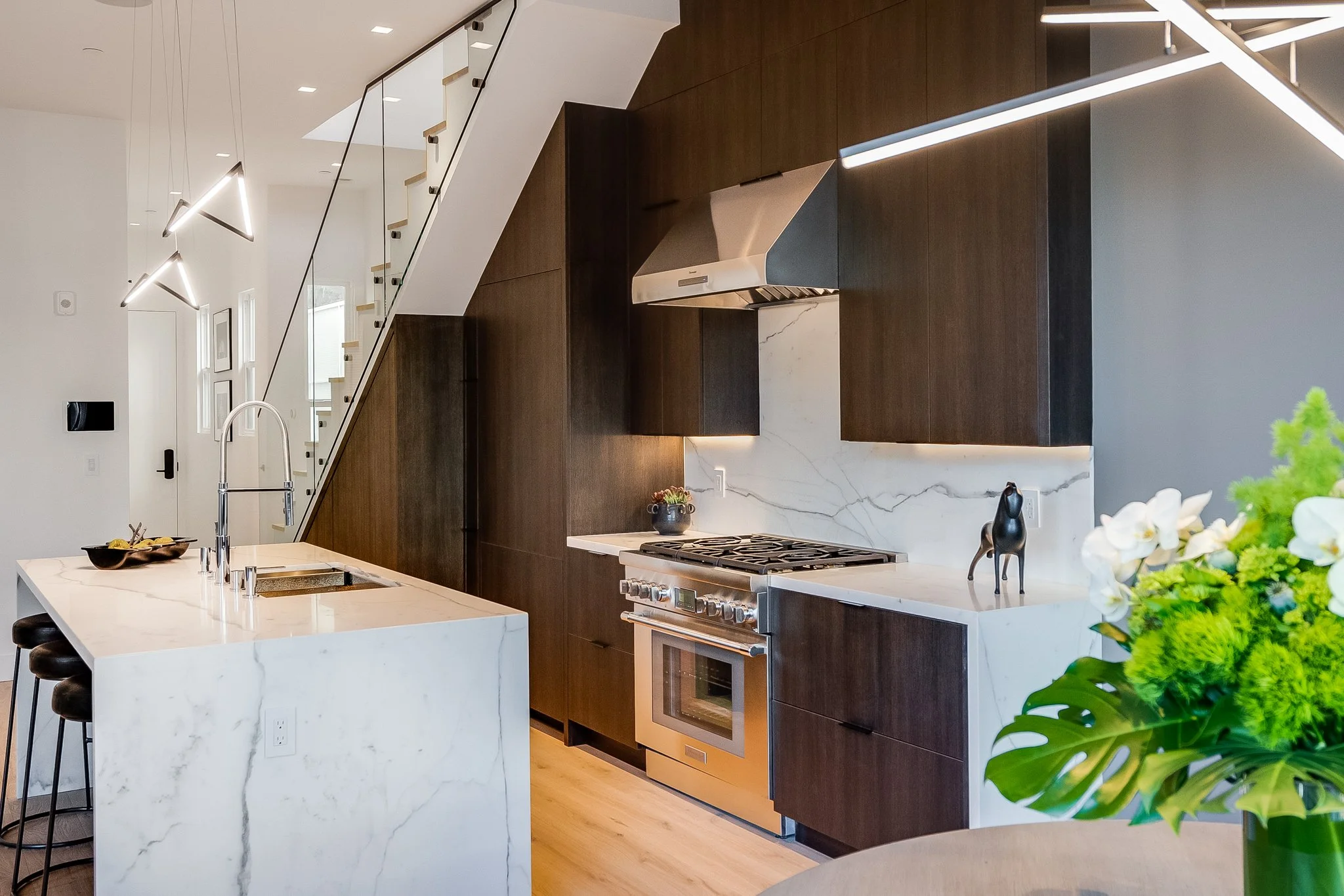 Modern kitchen with white marble island, dark wood cabinets, and stainless steel appliances. Features a staircase with a glass railing on the left and some decorative plants on the right.