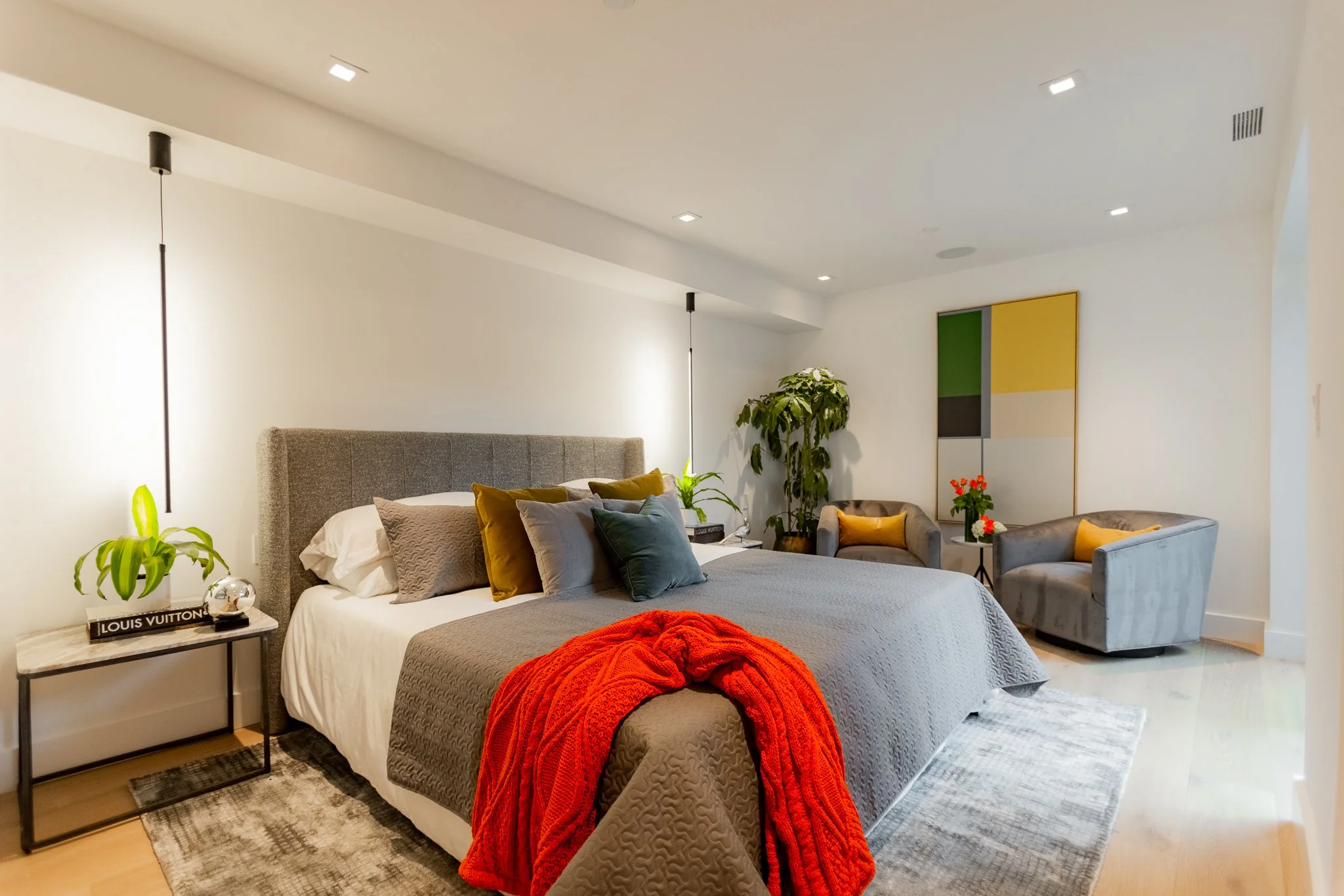 Modern bedroom with gray bed, colorful throw blanket, side table with plant, and seating area with two gray armchairs and a small table with flowers.