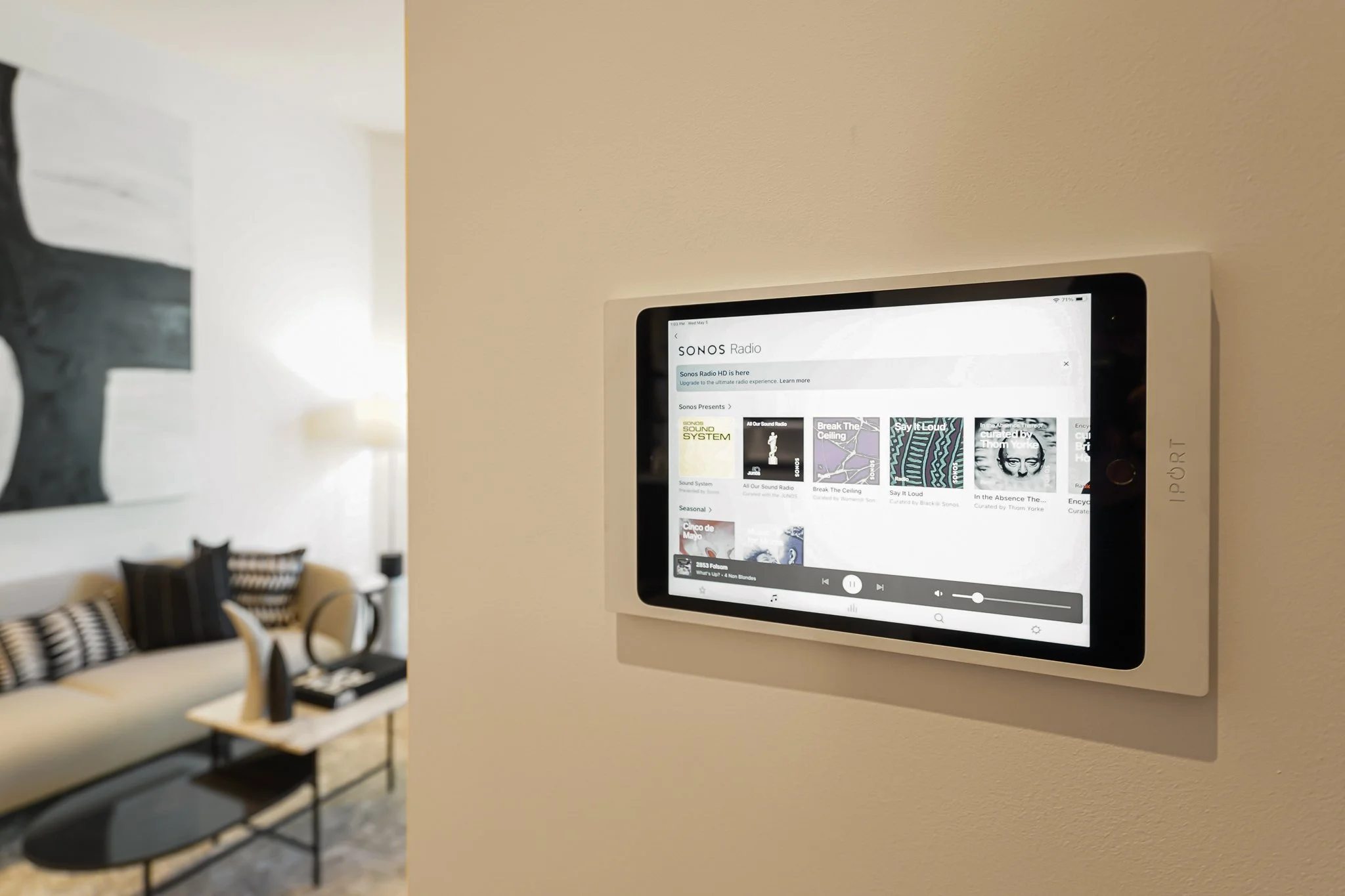 Wall-mounted digital display showing Sonos Radio app with playlists and album art in a living room with a sofa, pillows, a lamp, and a table.