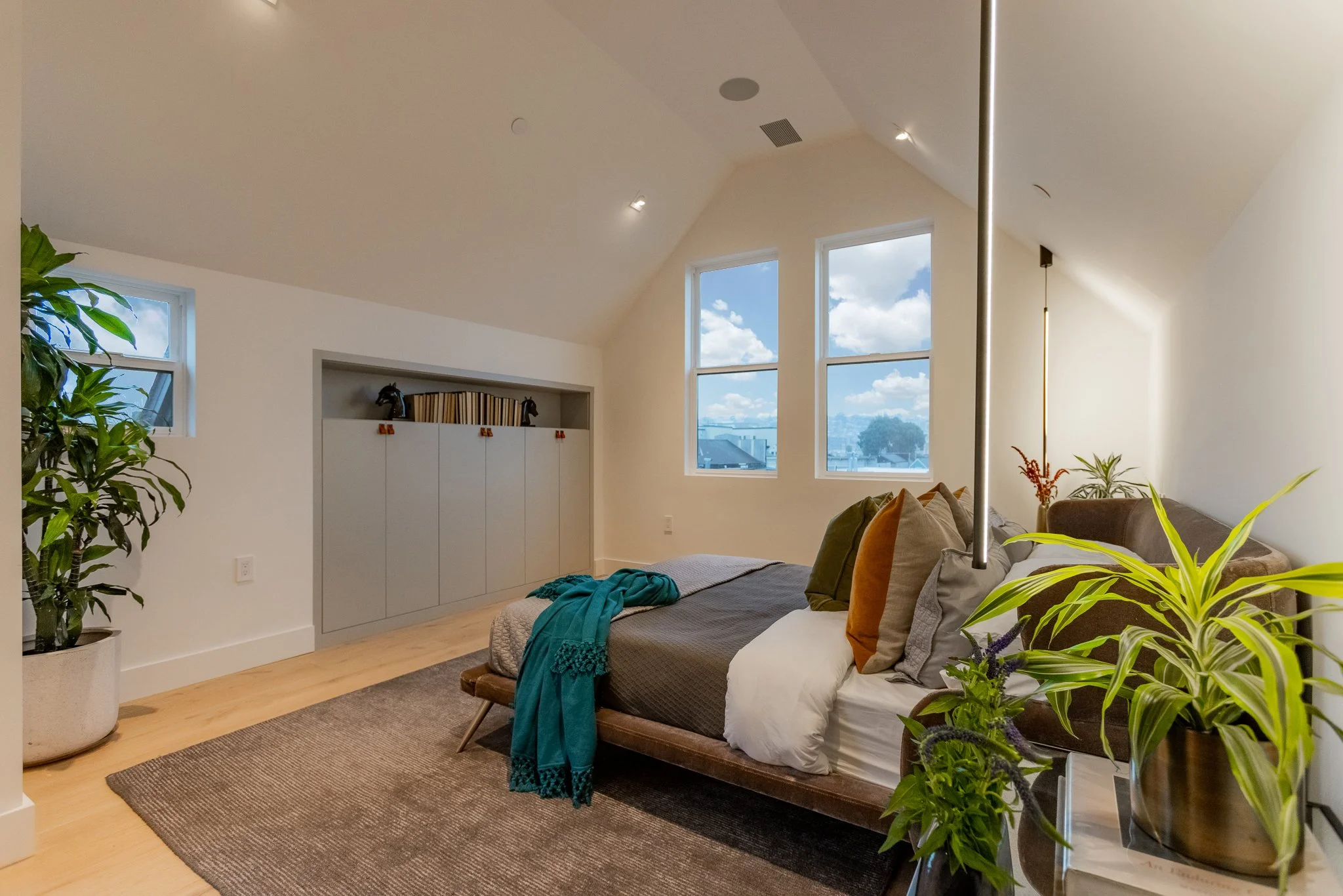 A modern bedroom with a sloped ceiling and three large windows showing a cityscape and blue sky with clouds. The room has a bed with multiple pillows, a throw blanket, and a teal scarf. There are several green plants, a brown rug, a built-in storage 