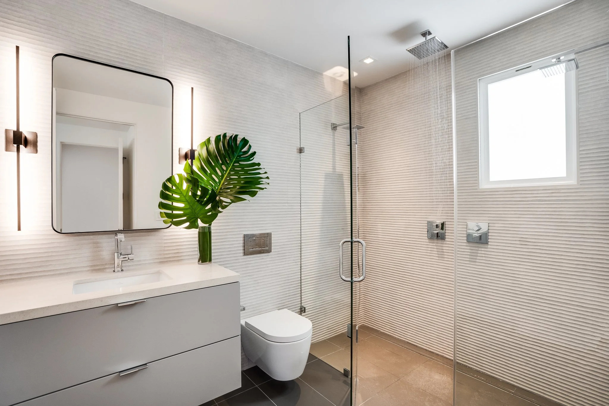 Modern bathroom with white textured walls, glass shower enclosure, large mirror, white vanity with a sink, a green plant in a glass vase, and a window letting in natural light.
