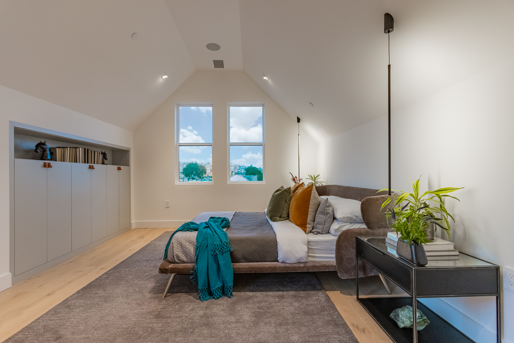 Modern bedroom with white walls, wooden flooring, two large windows showing a blue sky with clouds and trees outside, a bed with white linens, decorative pillows, and a teal blanket, a black side table with a plant and books, and a built-in storage c