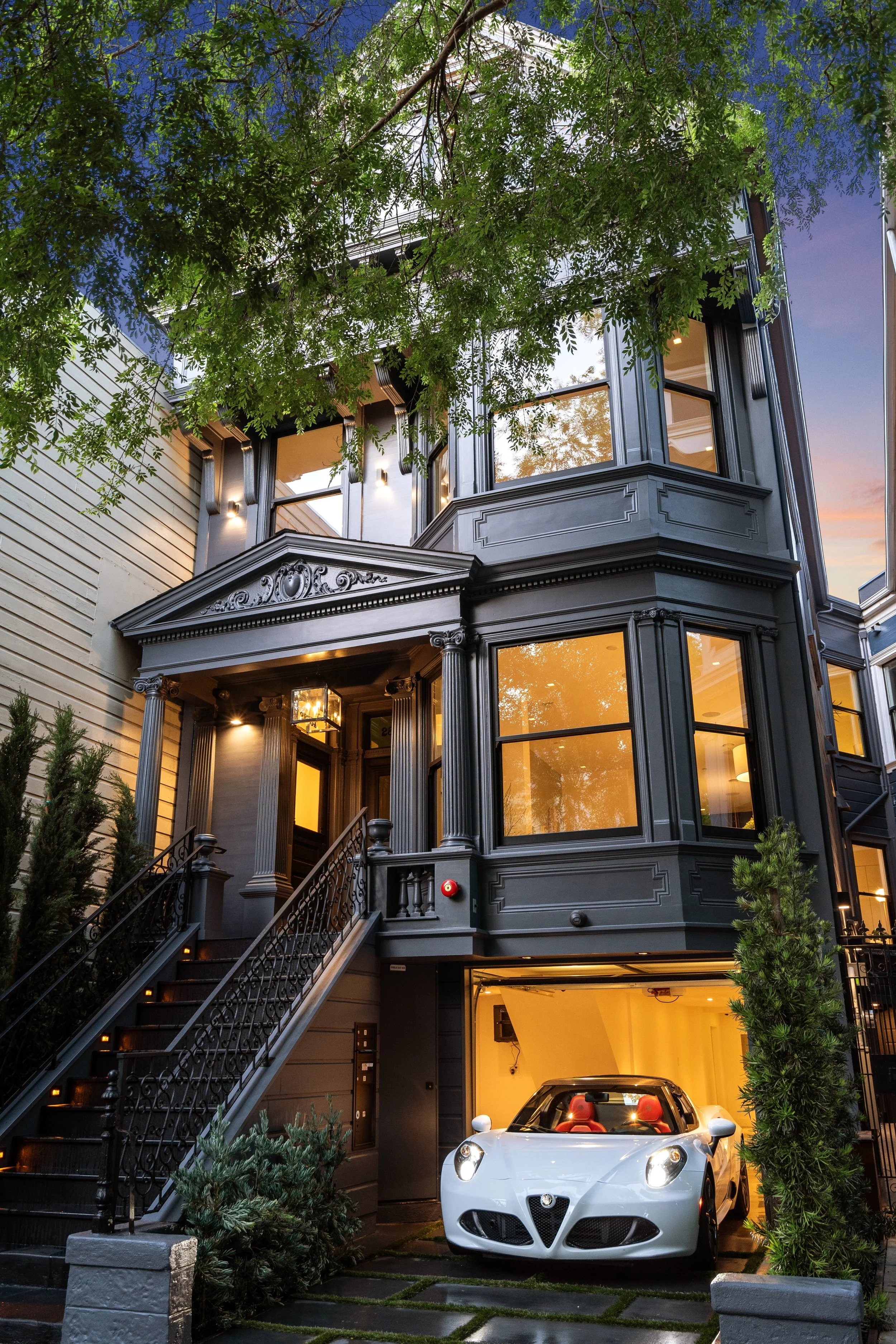 A multi-story Victorian-style house painted in black, with large windows, a staircase leading to a front porch, and a white sports car parked in a garage beneath the house. There are trees and greenery around the house, and the sky is at sunset.