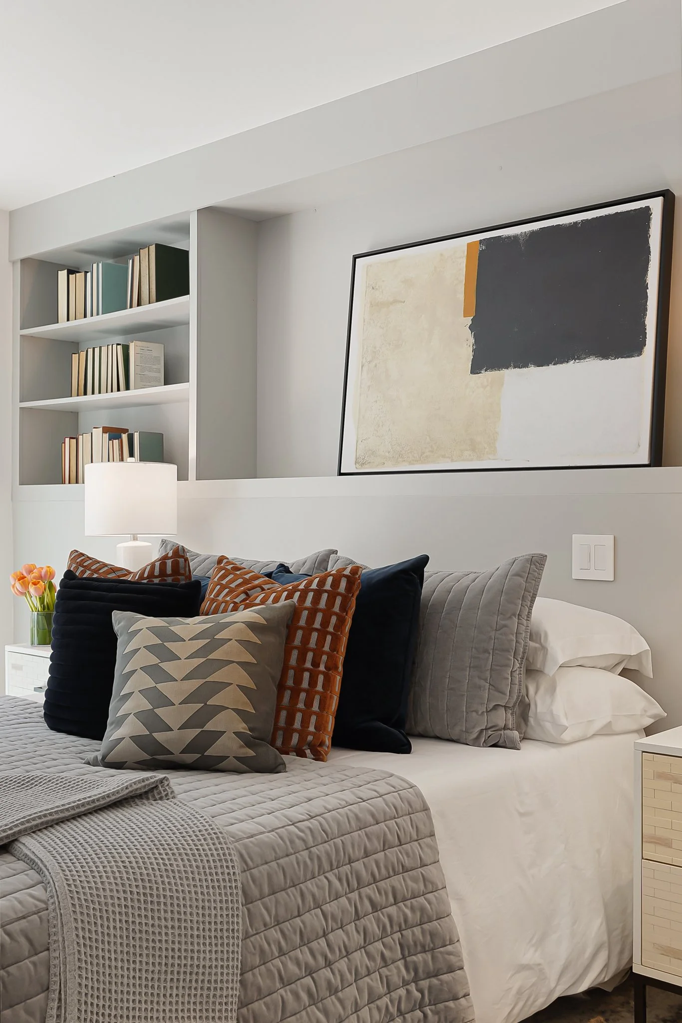 A cozy bedroom with a bed covered in a light beige quilt, topped with numerous decorative pillows in black, gray, and orange-brown patterns. There is a bedside table with a lamp and a vase of pink tulips. Above the bed, abstract artwork in black, bei