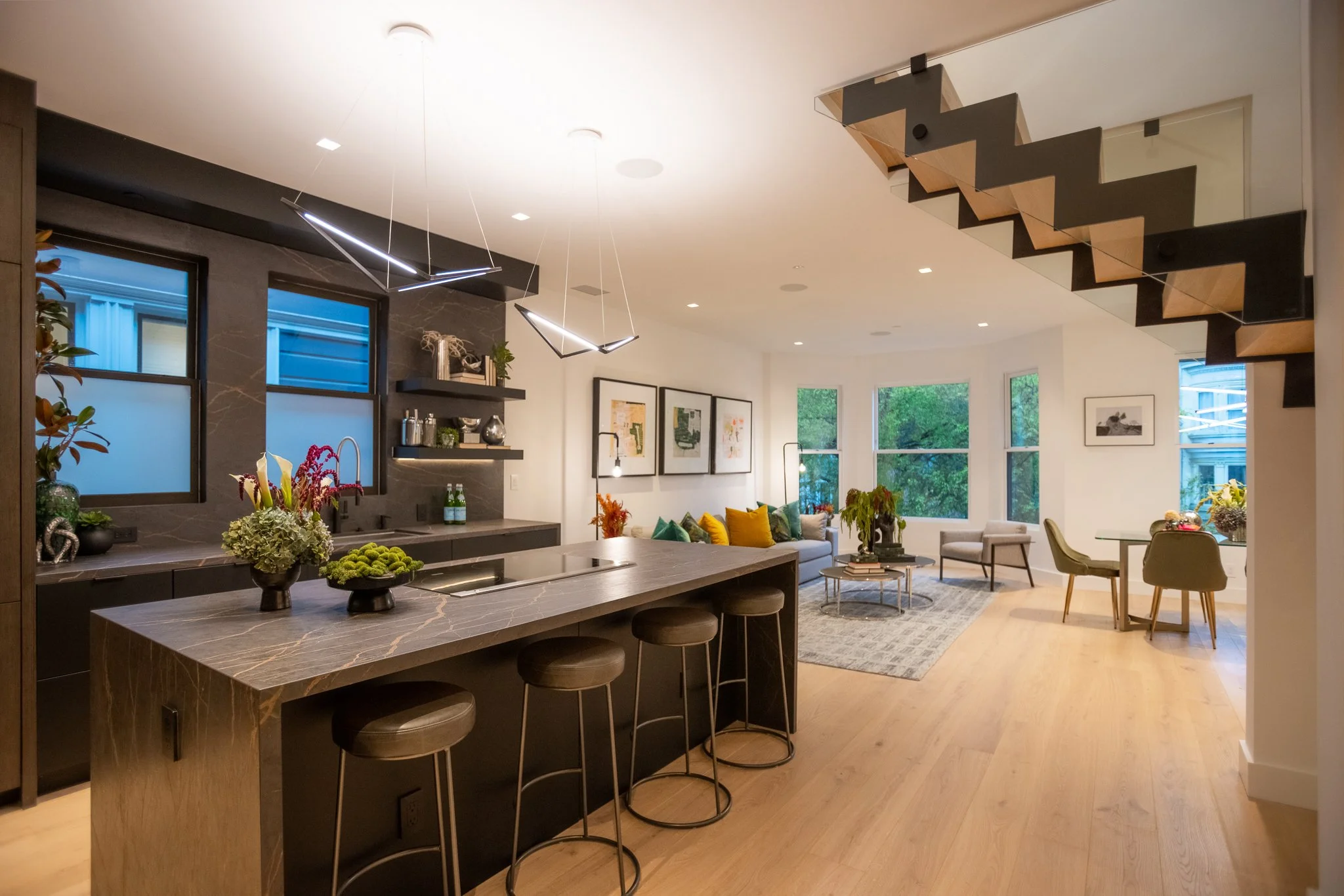 Open-concept living room and kitchen with staircase, bar stools, and large windows showing greenery outside.