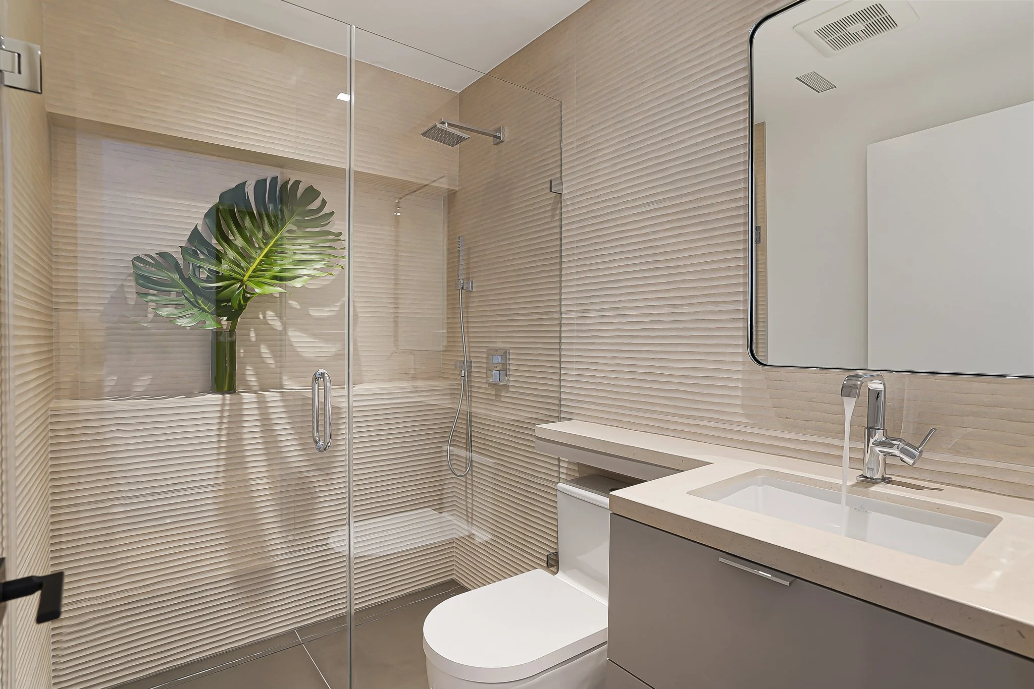 Modern bathroom with beige textured walls, a glass-enclosed shower with a large green leaf plant in a vase, a white toilet, and a vanity with a sink and mirror.