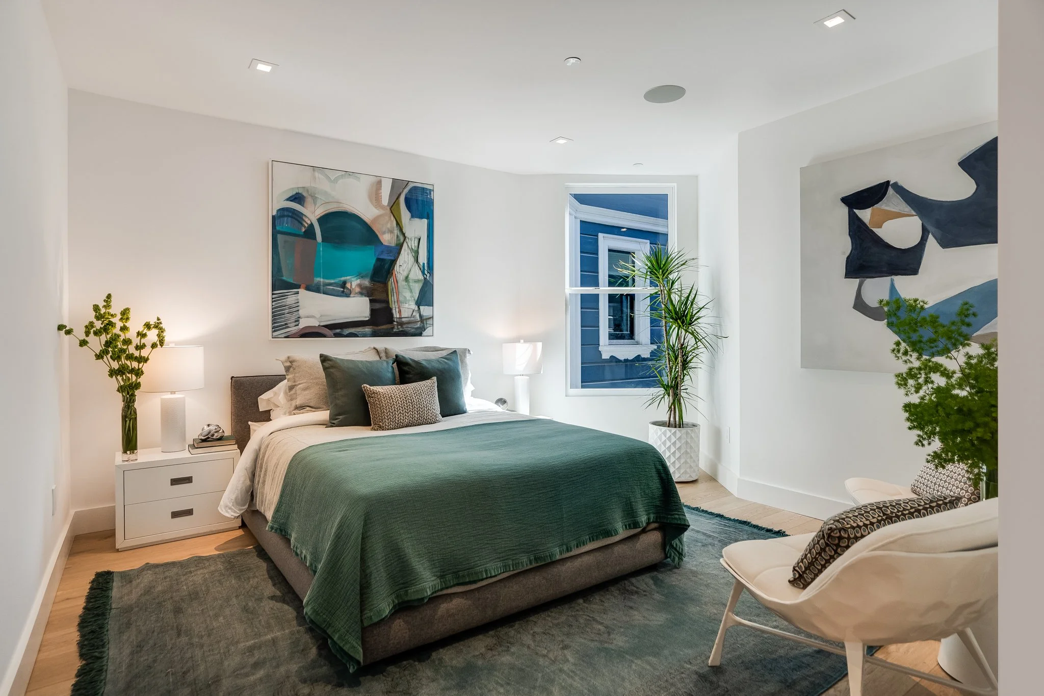 Modern bedroom with green bedding, white walls, abstract art on the wall, a potted plant near the window, and a white chair with patterned pillow.