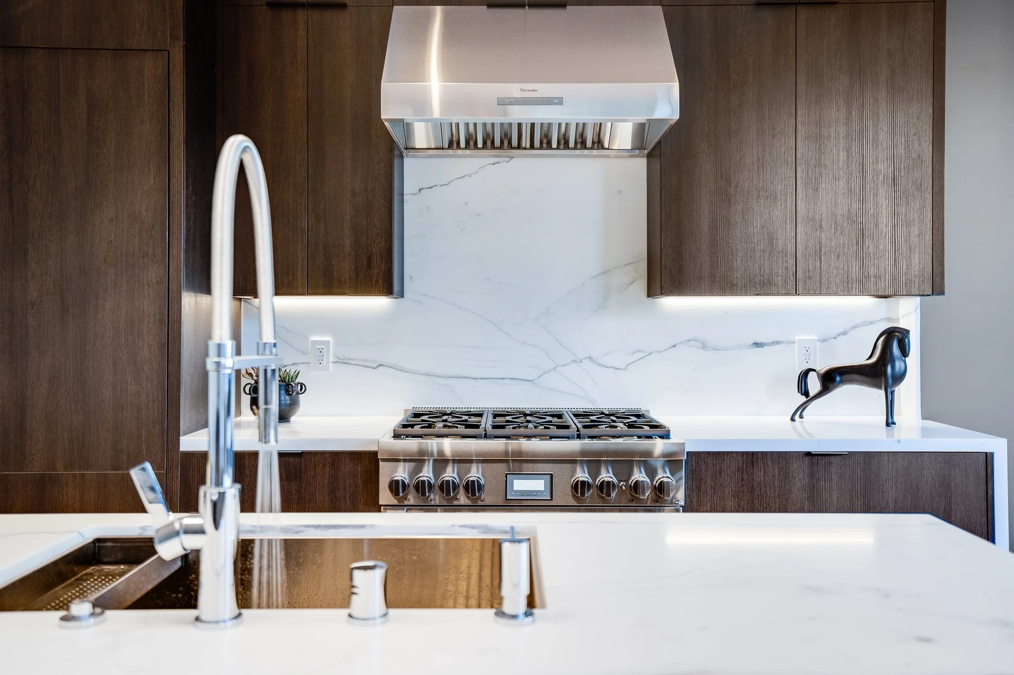 Modern kitchen with white marble backsplash and countertops, dark wood cabinets, and stainless steel appliances including a stove and range hood. Decor includes a small plant and a black dog sculpture.