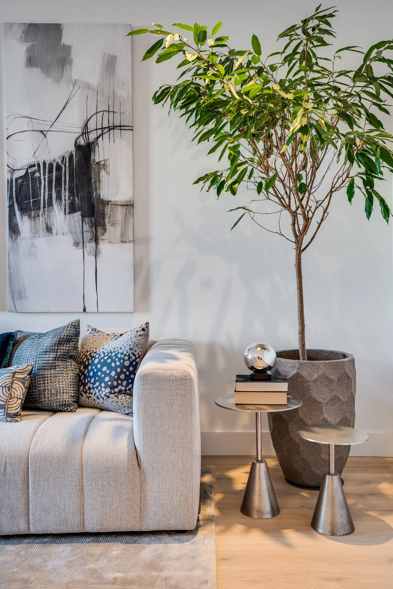 Living room with a beige sofa, patterned pillows, large potted tree, abstract black and white painting, two metallic side tables, a reflective decorative sphere, and a small stack of books.