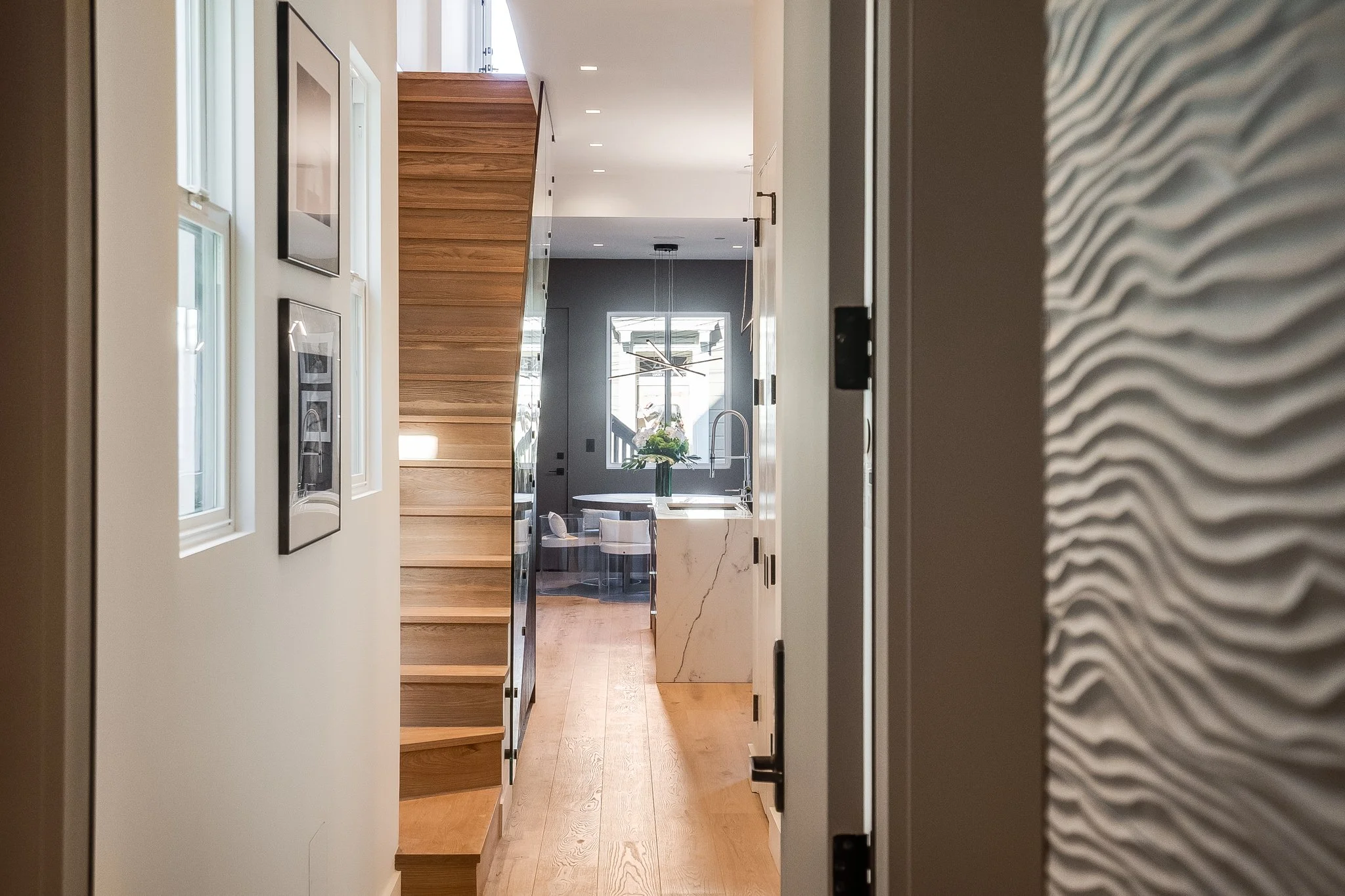 A modern hallway leading to a kitchen, featuring wooden stairs on the left, a marble island with a vase of flowers, and large windows in the background.