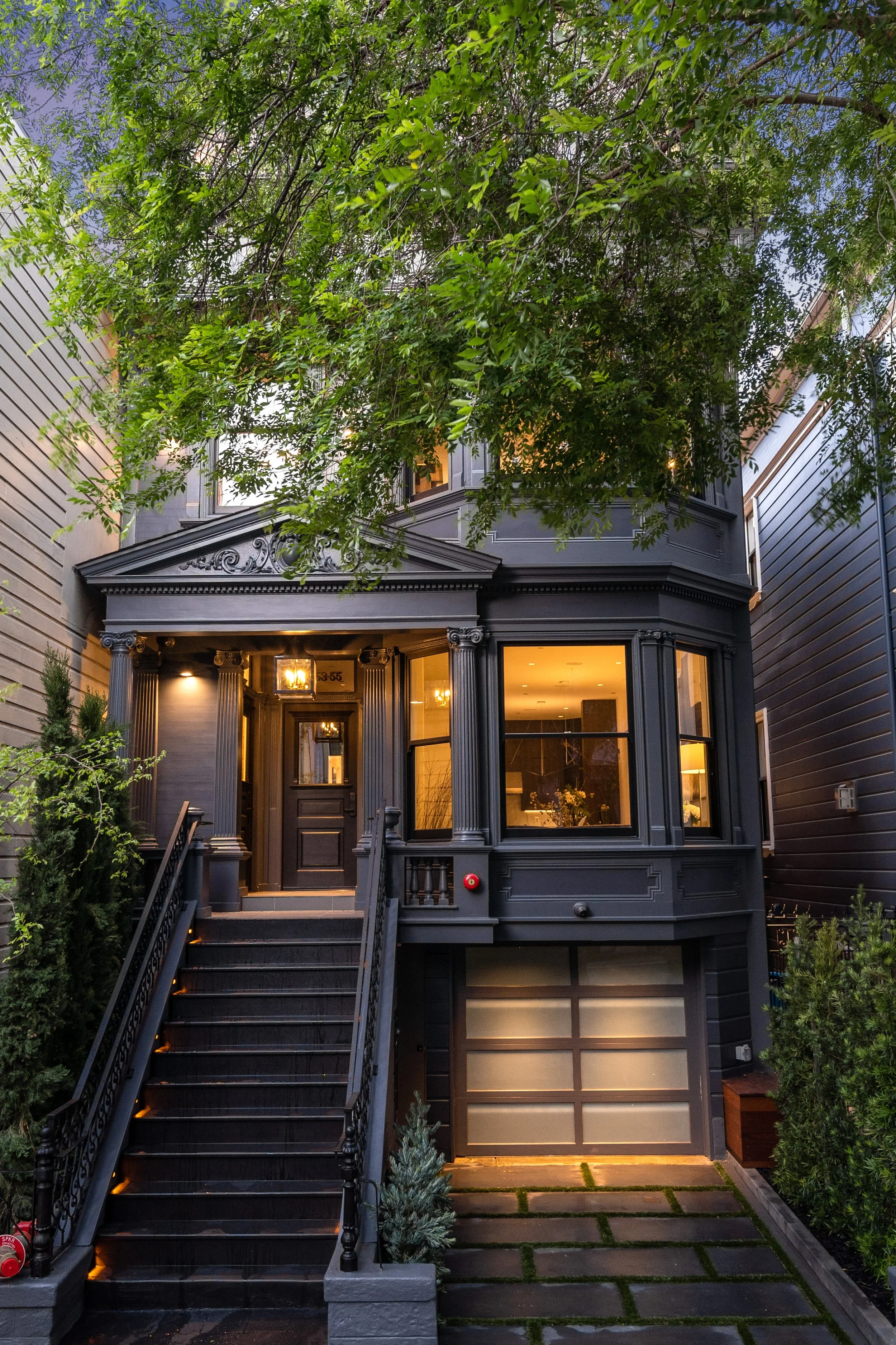 A black, multi-story house with a front staircase, bay windows, and decorative columns, surrounded by greenery and trees.
