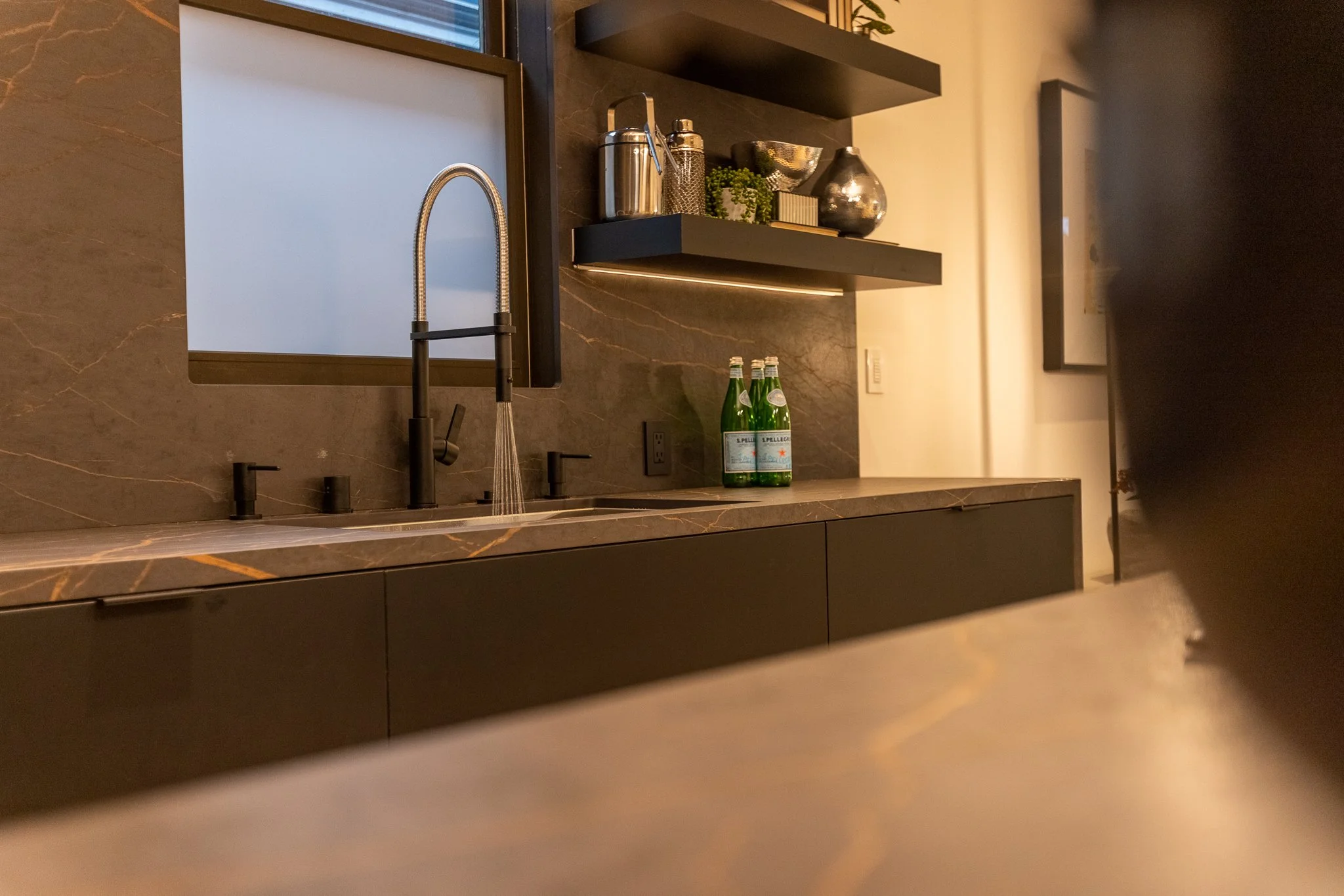 Modern kitchen with black countertops, a sleek black sink with a high curved faucet, two bottles of San Pellegrino sparkling water on the counter, open black shelves with various metallic and decorative items, and a window with frosted glass.