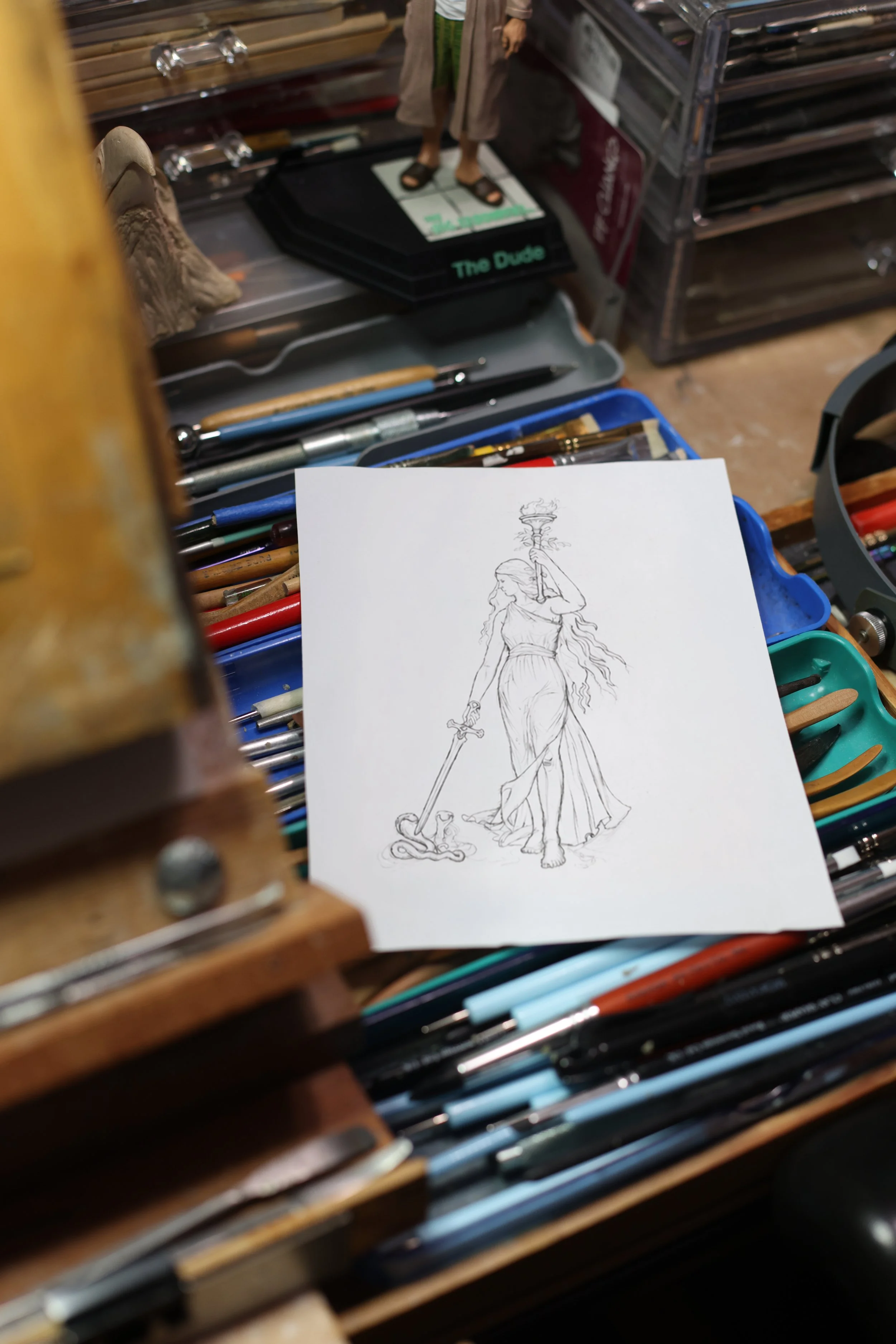 Concept sketch for a coin or medal design displayed on an artist's workbench surrounded by professional drawing and sculpting tools. The image showcases the creative process behind numismatic art, from initial illustration to finished coin design.
