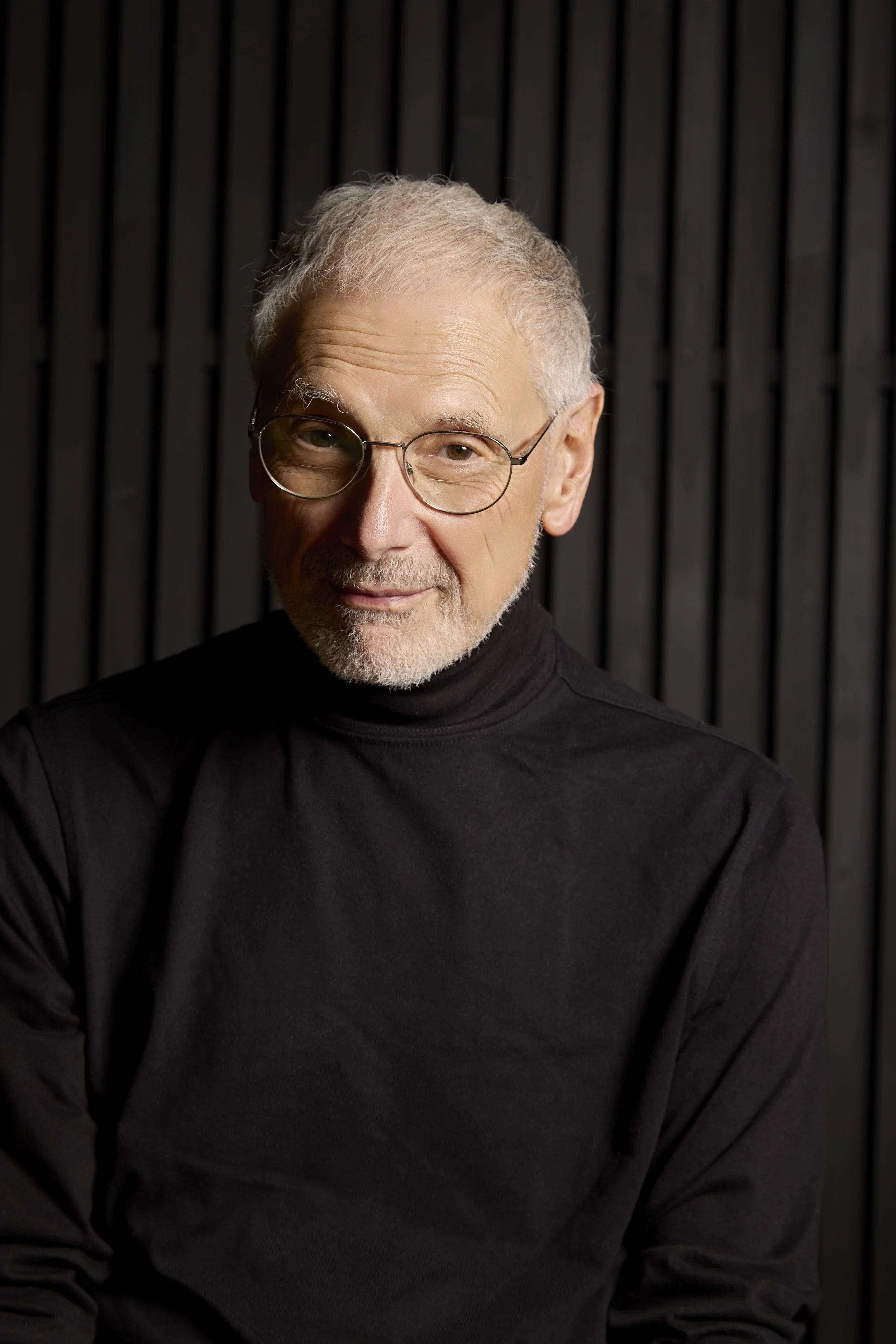 A close-up portrait of an elderly man with gray hair, glasses, and a beard, wearing a black turtleneck, against a dark, vertical striped background.