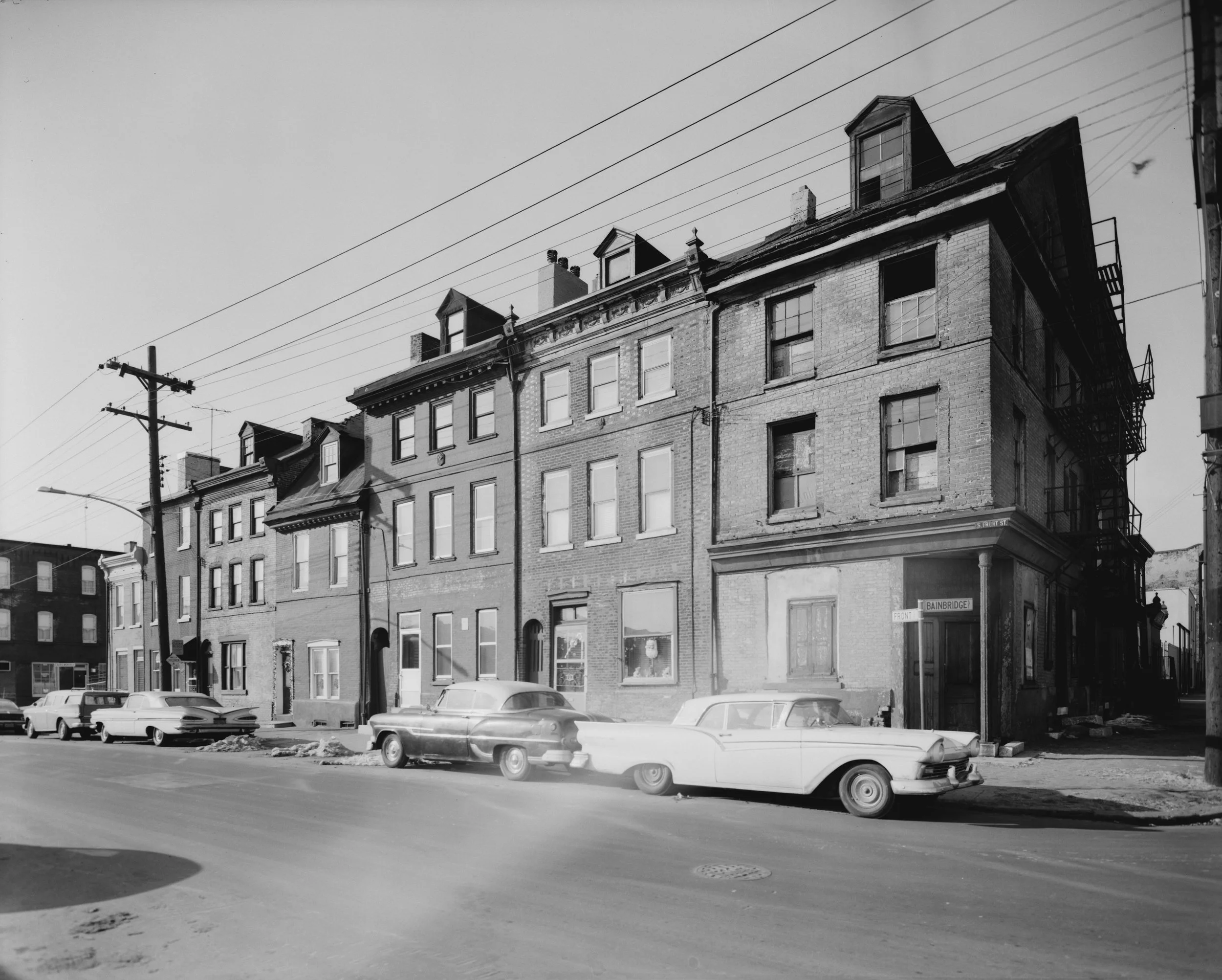 Historical black-and-white photograph of a South Philadelphia street lined with brick row houses and parked vintage cars, displayed beside text about John Mercanti's early years in the 1940s.