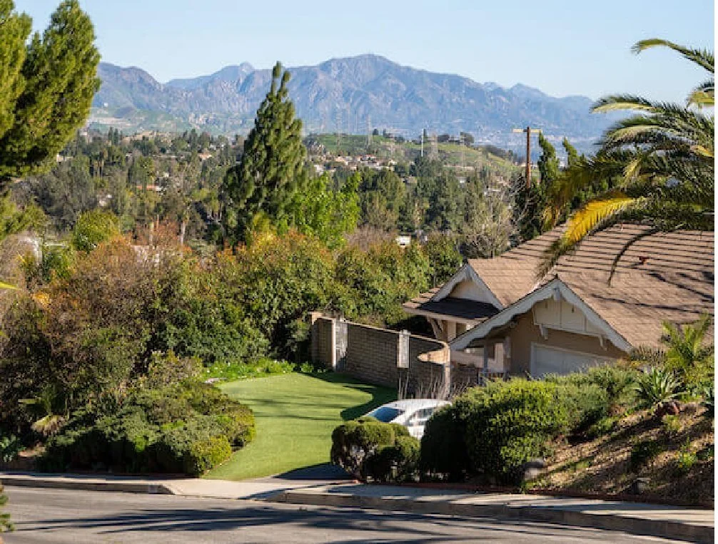 Residential neighborhood with a house, well-maintained lawn, trees, and mountains in the background under clear blue sky.