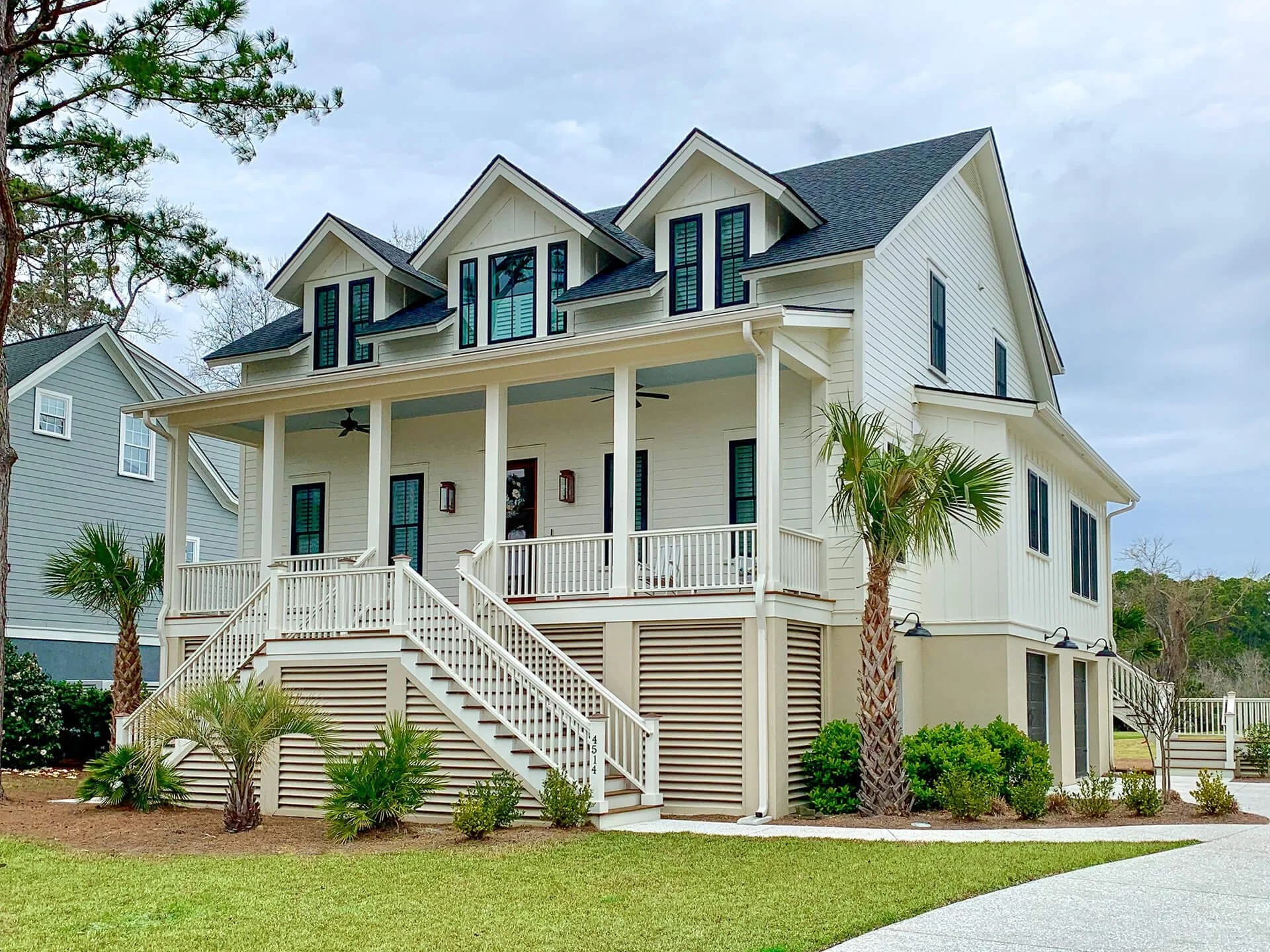 A three-story detached house with white exterior and black windows, front porch with white railings, stairs, and decorative palm trees in the yard, under a cloudy sky.