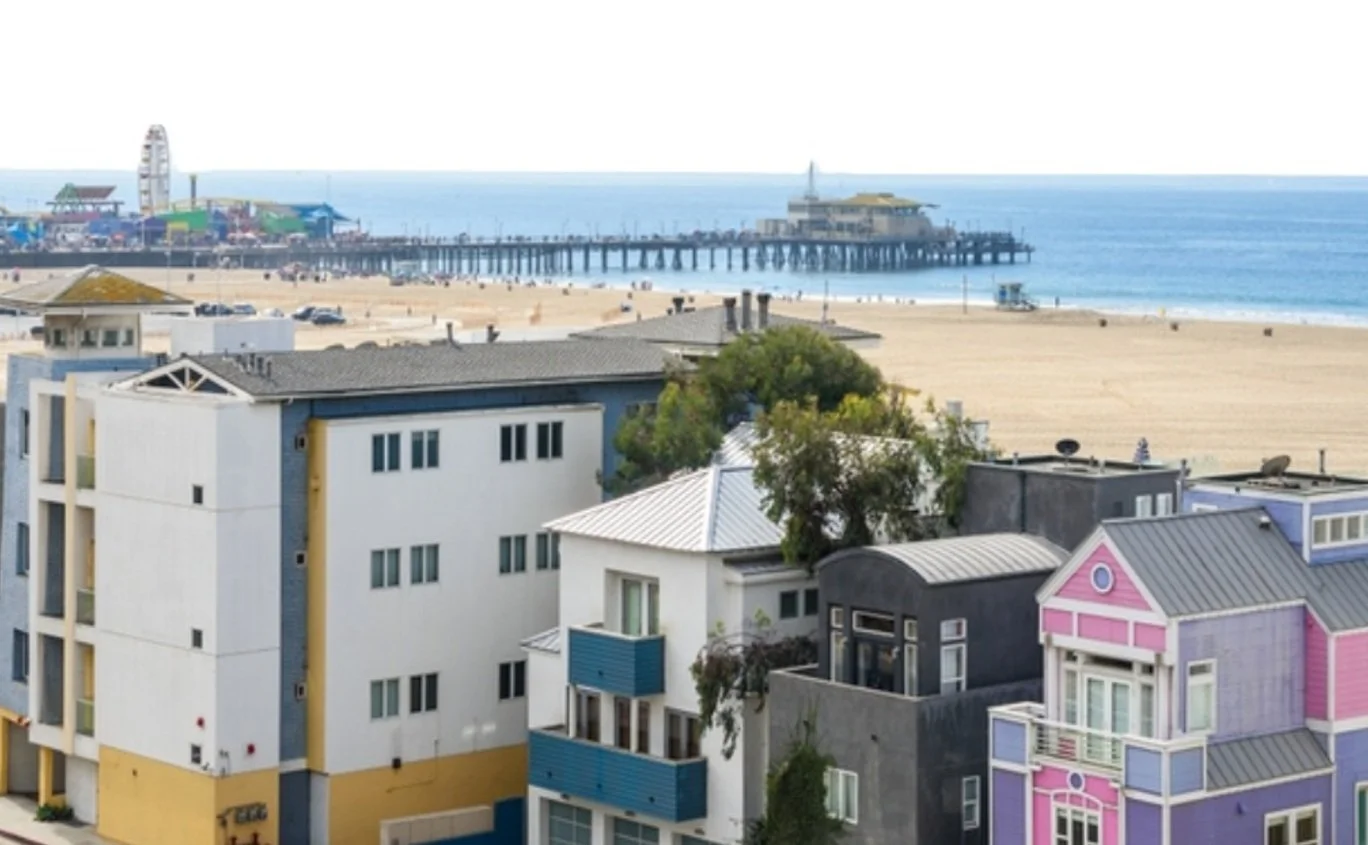 View of a beach with colorful houses in the foreground and a pier extending into the ocean with a Ferris wheel and amusement park in the background.