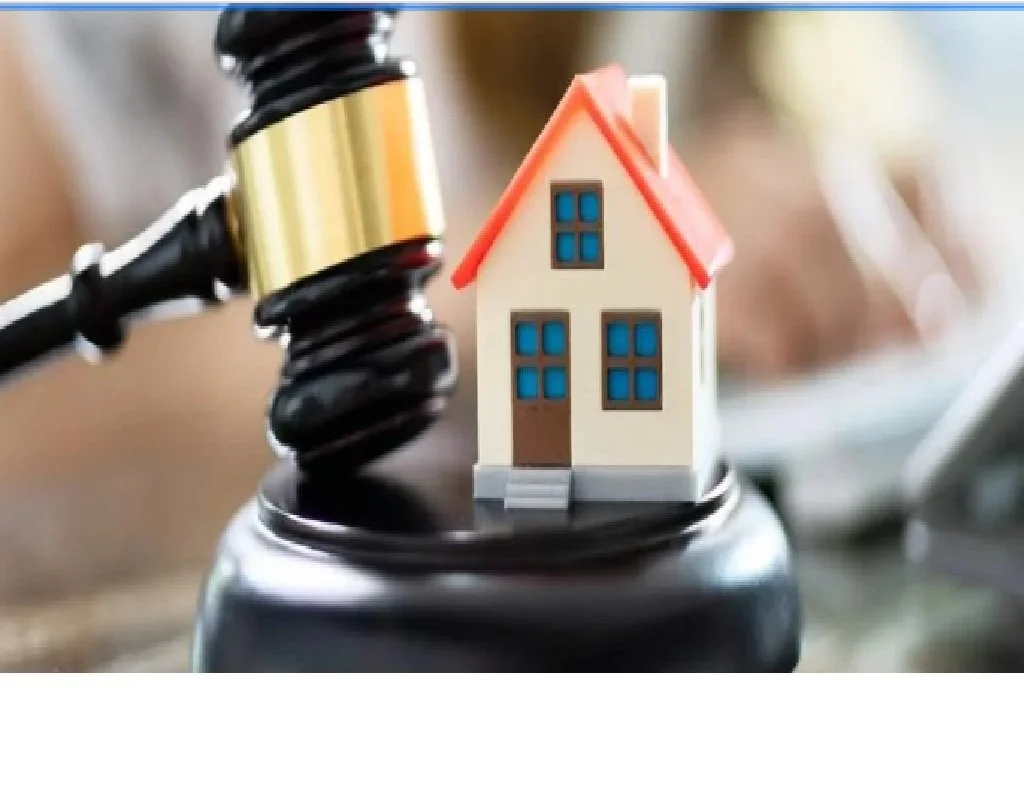 A miniature house model with blue windows and a red roof, placed on a black cause or loudspeaker, with a gavel in the background.