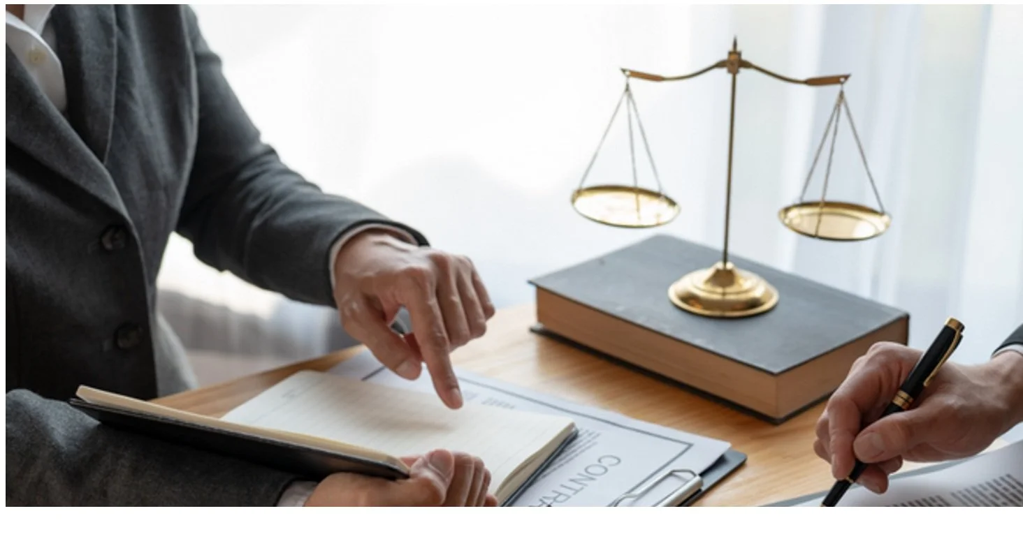 Two people are seated at a table, reviewing documents. One person is pointing at a notebook, while the other is holding a pen. On the table, there is a book, a contract, and a golden balance scale, with light coming from a window behind.