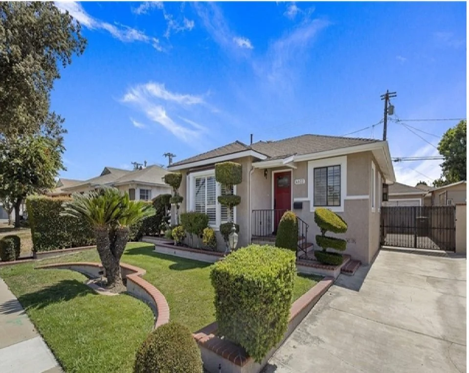 Front yard of a single-story house with manicured bushes, trees, and a well-maintained lawn. The house has beige exterior walls, white window frames, and a red front door. There is a sidewalk in front and a driveway to the side, with a black gate in the background under a bright blue sky.