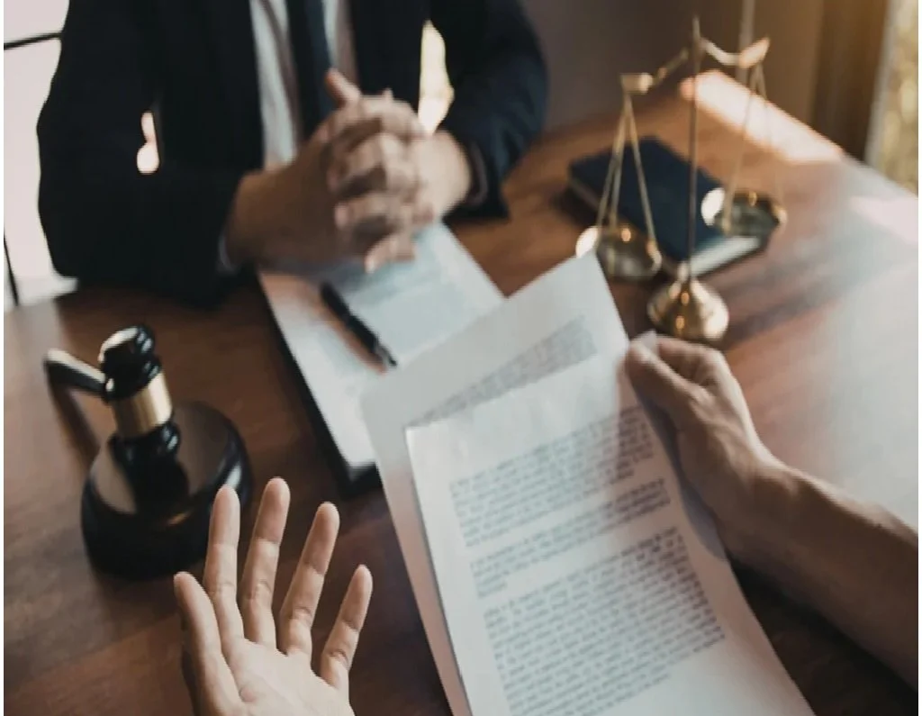 A person in a suit and another person with a watch on their wrist are exchanging documents at a wooden table, which also holds a brass scale, a black notebook, and a gold and black ink bottle.