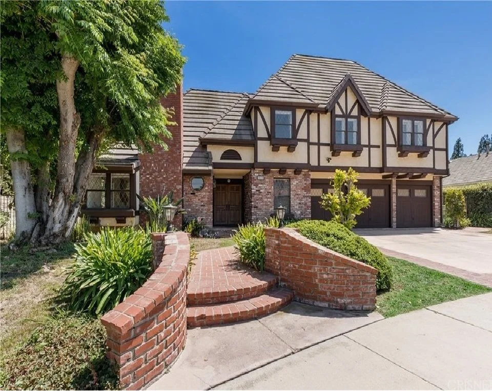 A two-story house with a brick and Tudor-style exterior, a three-car garage, a brick front walkway with curved steps, surrounded by green shrubbery and a large tree, under a clear blue sky.