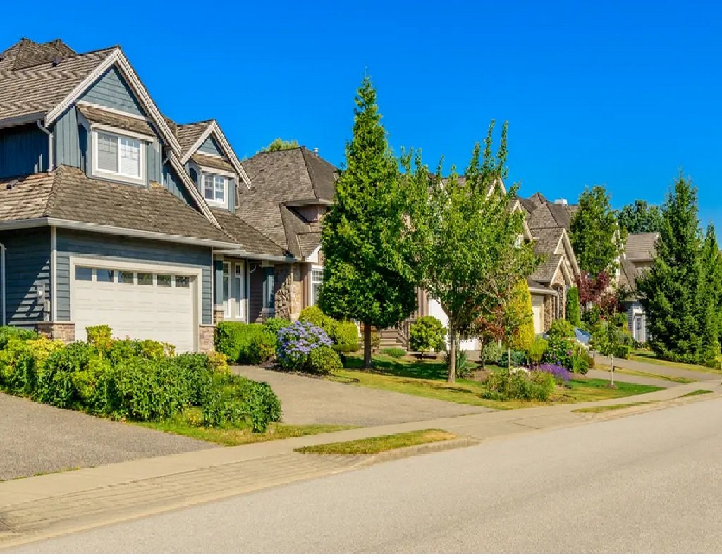 Neighborhood street with modern houses, well-maintained lawns, and trees under a clear blue sky.