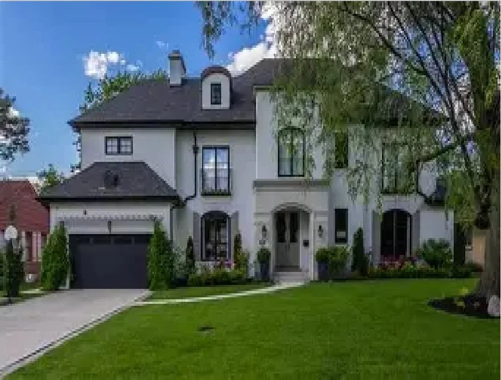 A modern, white two-story house with a dark roof, black garage door, and front porch. The house has tall, narrow windows and is surrounded by a green lawn with trees and shrubs.