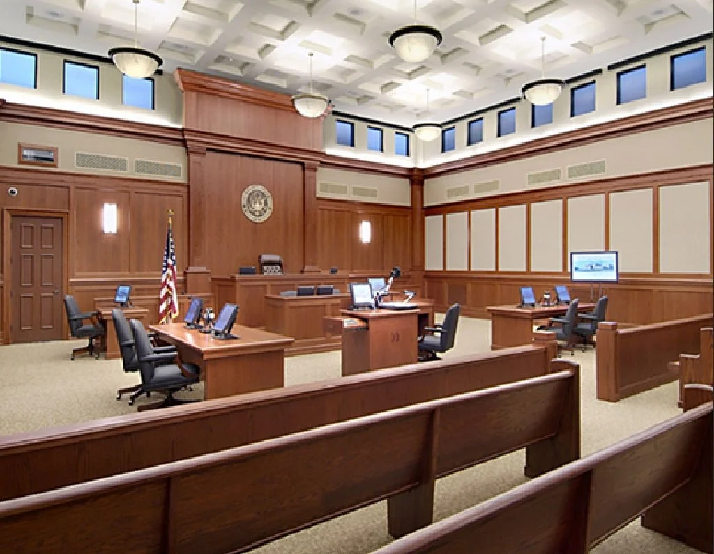 A courtroom with wooden furniture, a judge's bench, court clerk's desk, and multiple chairs, with digital screens on desks and an American flag in the corner.