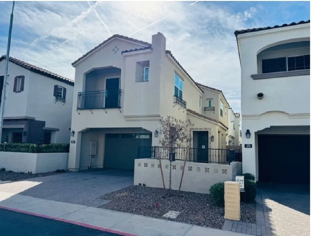 A row of modern, two-story houses with white exteriors and black roofs. The house in the center has a small balcony and a garage, with a small landscaped front yard and a young tree. The neighboring houses also have garages and similar designs, with a clear blue sky overhead.
