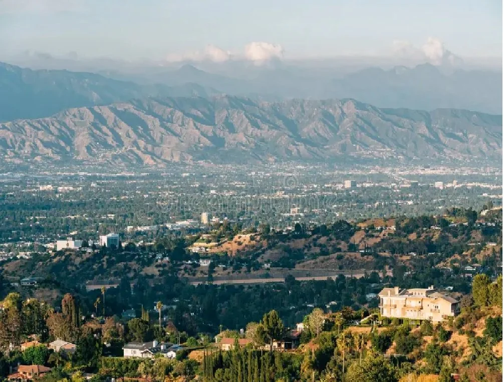 View of a city with natural landscape, featuring mountains in the background, and residential houses and trees in the foreground.