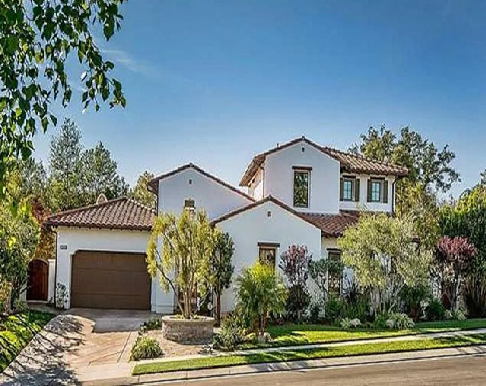 A two-story house with a white exterior, brown tile roof, attached garage, and landscaped front yard with trees and bushes.