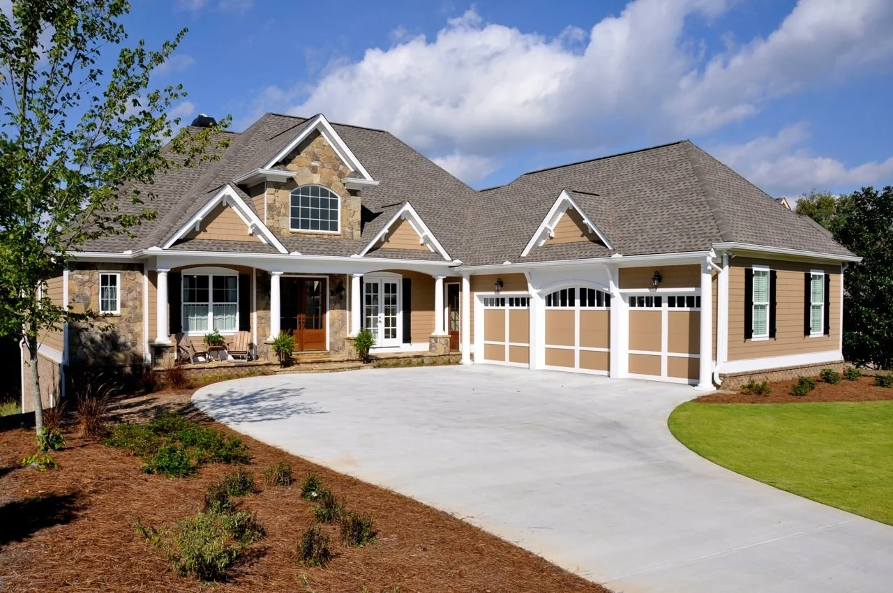 Front view of large suburban house with stone and beige siding, three-car garage, manicured lawn, and a partly cloudy sky.
