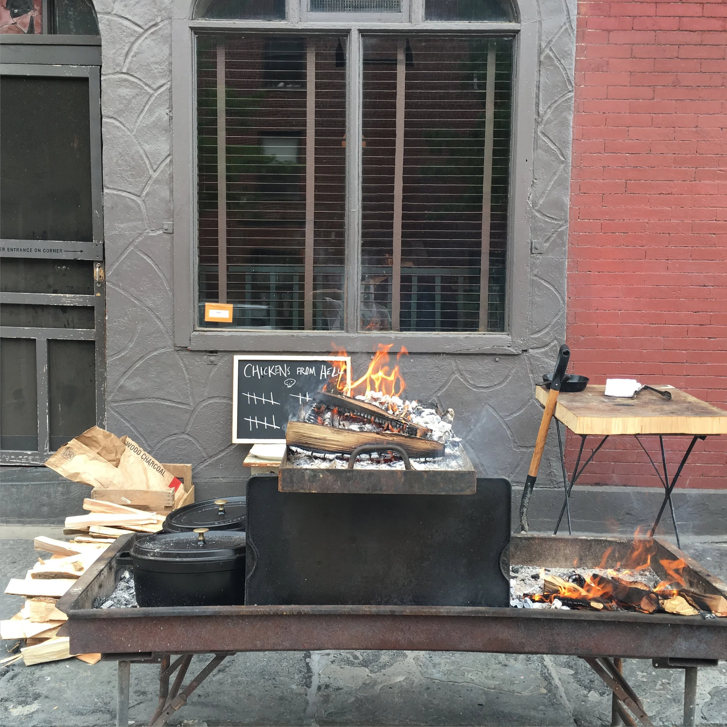 A wood-fired smoker grill with open flames, wood logs inside, and two black cast iron pots on a lower shelf. To the right, there is a small wooden table with a meat thermometer and a white paper towel. To the left, a brown paper bag and wood pieces a