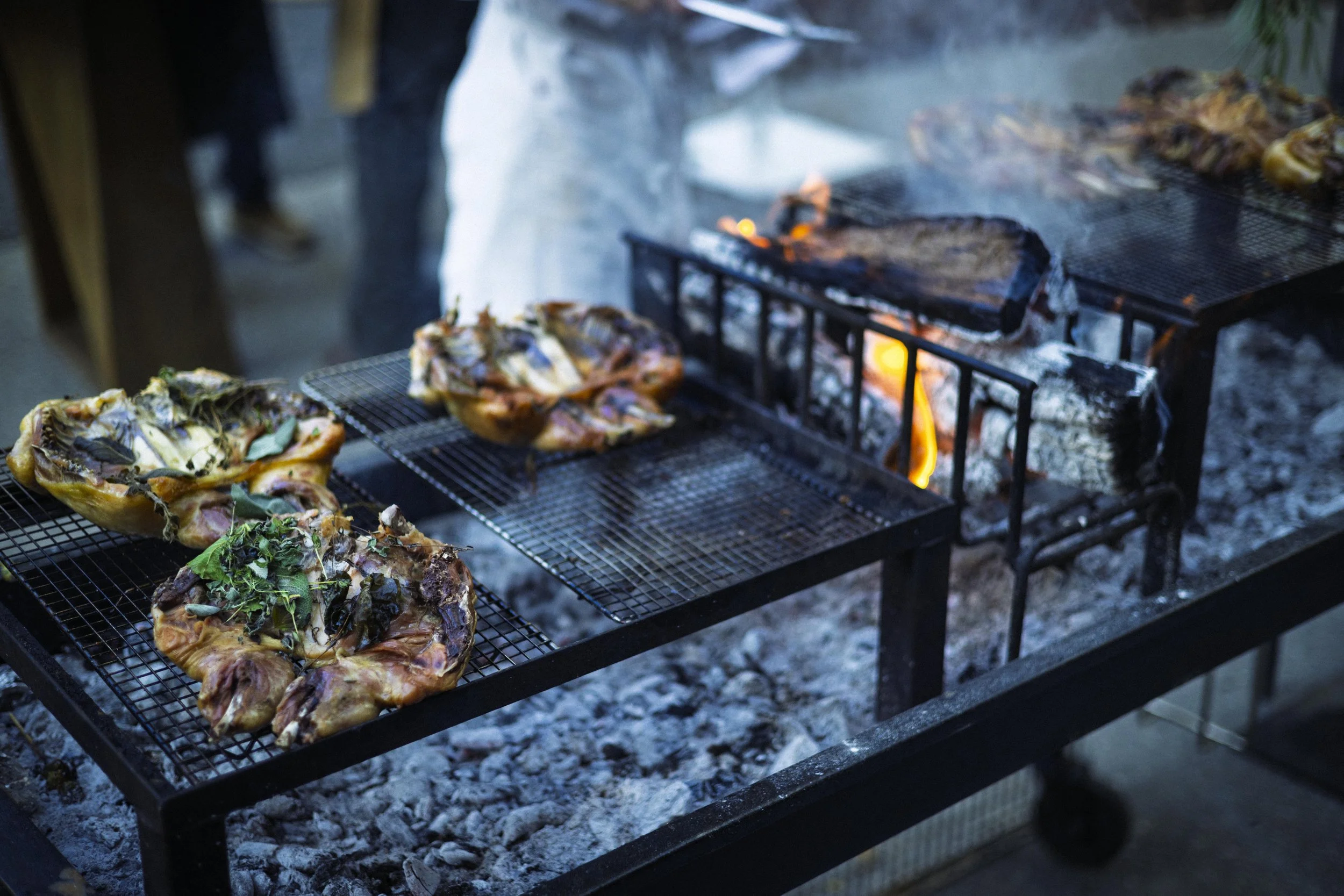 Open outdoor grill with several pieces of meat and vegetables cooking over charcoal, flames visible underneath.