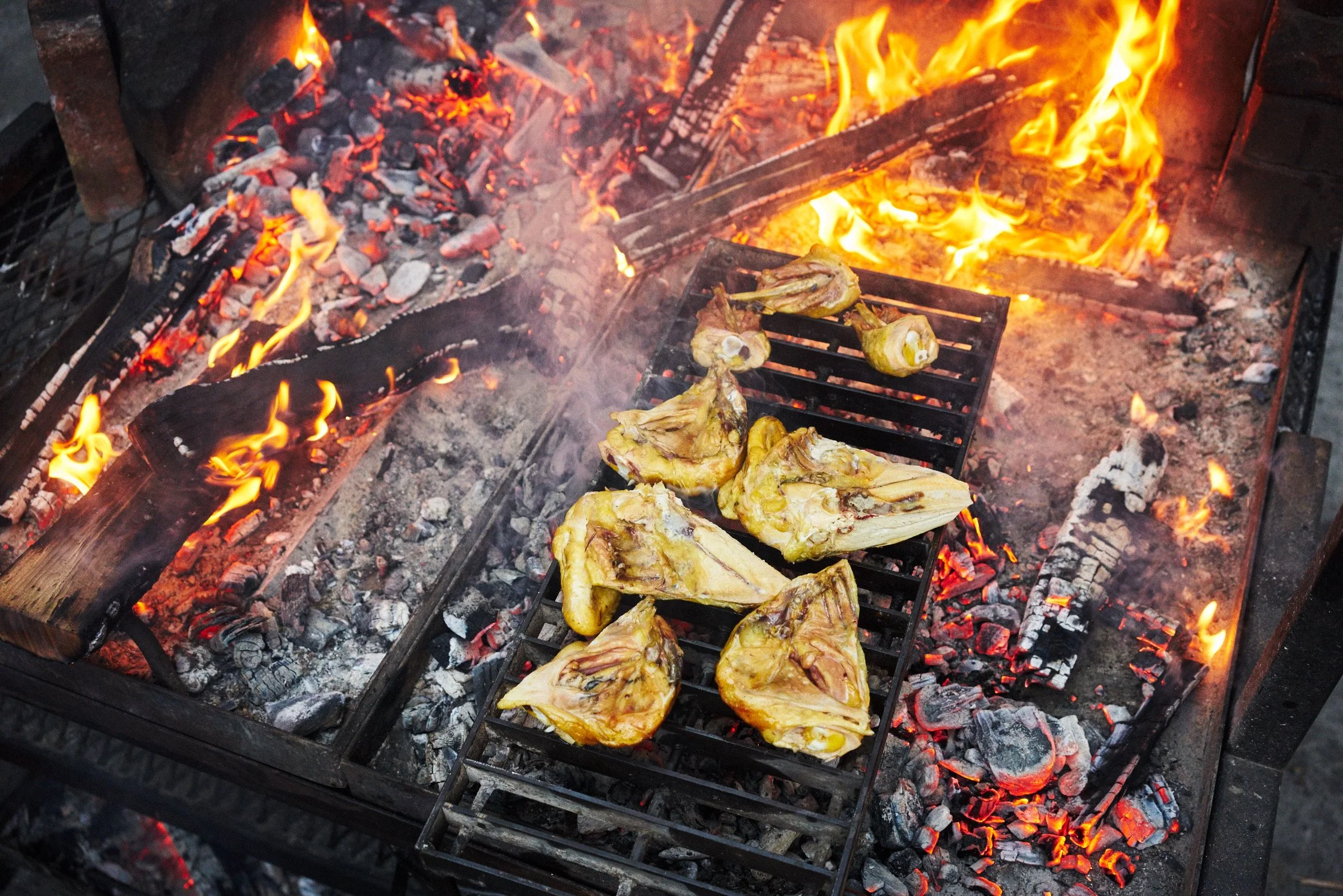 Pieces of chicken grilling over an open fire with glowing hot coals and flames.