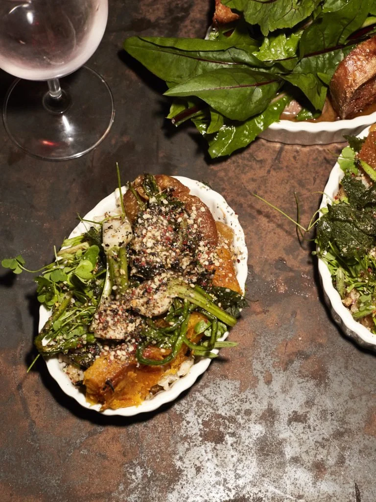 A white dish filled with roasted vegetables, garnished with greens and sprinkled with cheese, on a dark table alongside a glass of white wine.